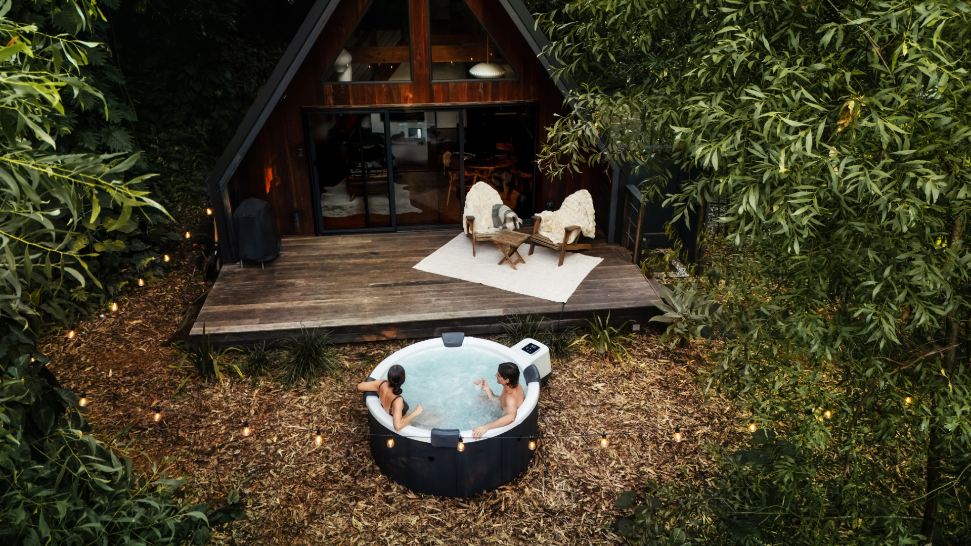 Zwei Personen in einem Whirlpool. A-Rahmen-Hütte im Hintergrund mit Stühlen auf dem Deck. Bäume umgeben die Szene, mit Lichterketten.