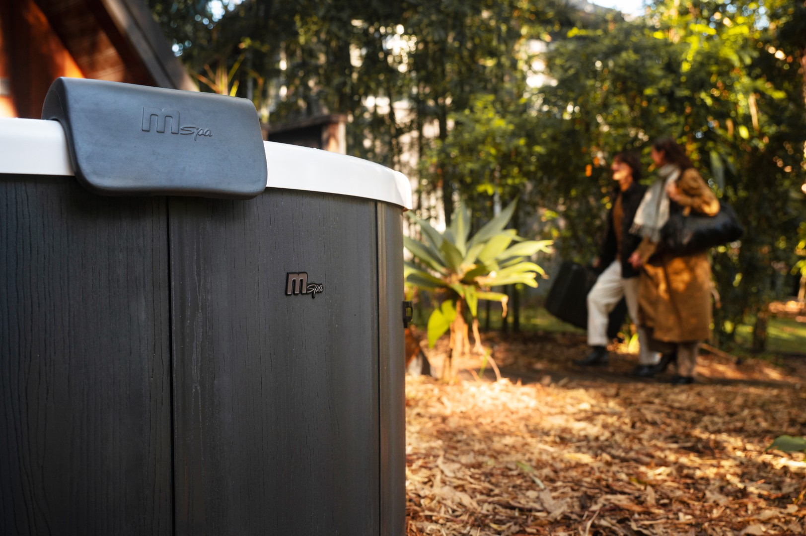 Ein dunkler Whirlpool mit dem Logo 'mSpa' in einer Gartenumgebung. Zwei Personen gehen im Hintergrund.