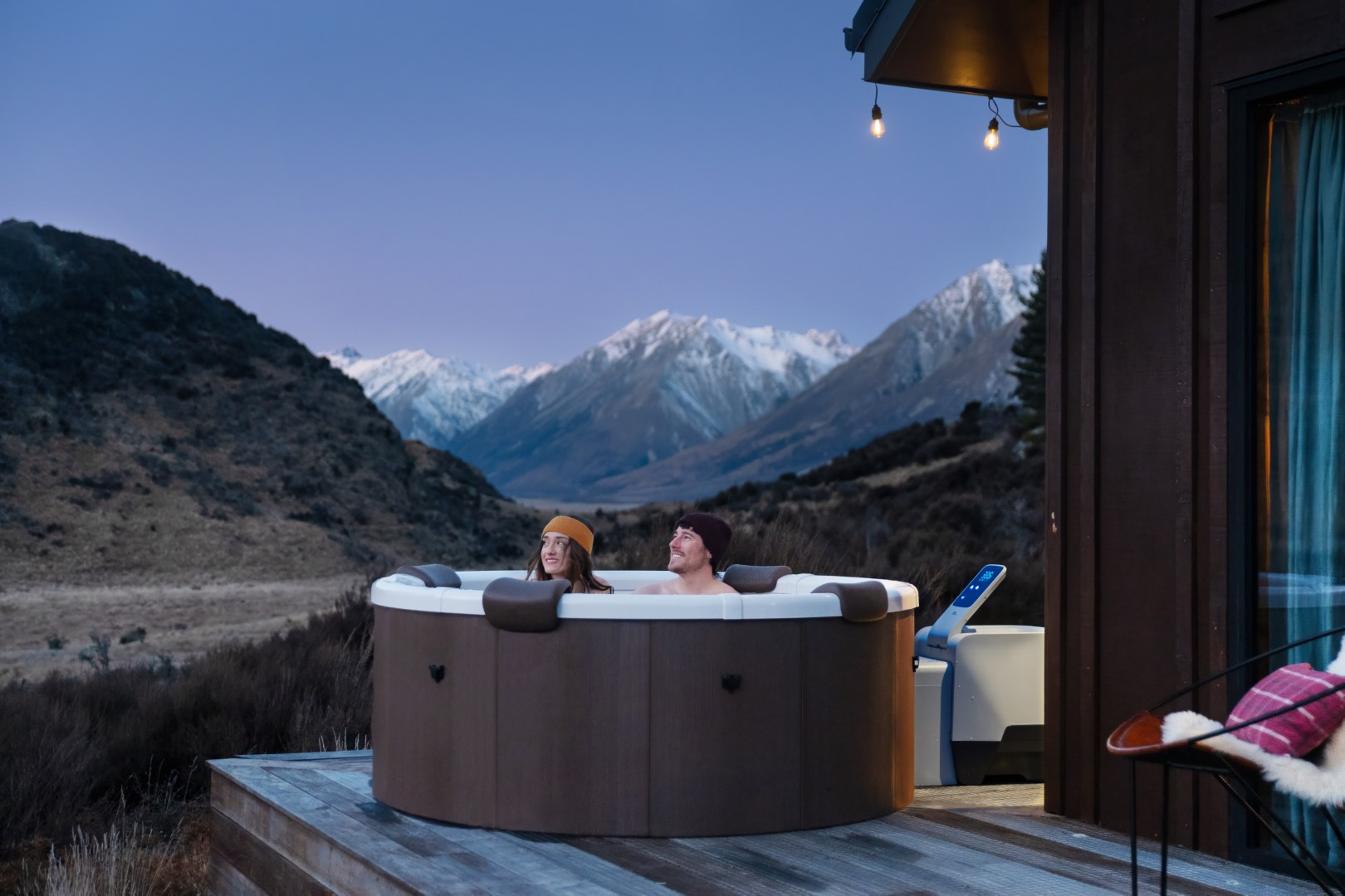 Ein Paar entspannt sich in einem Whirlpool im Freien, im Hintergrund Berge. Der Whirlpool steht auf einer Holzterrasse.