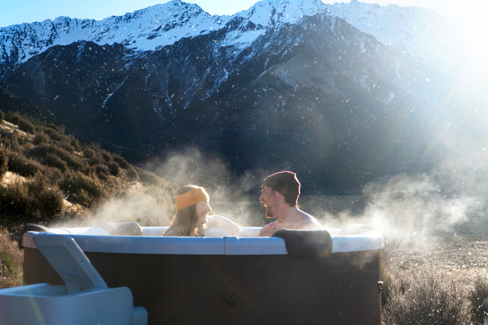 Ein Paar genießt einen Whirlpool mit Bergblick. Dampf steigt auf. Schneebedeckte Berge im Hintergrund. Klarer Himmel.