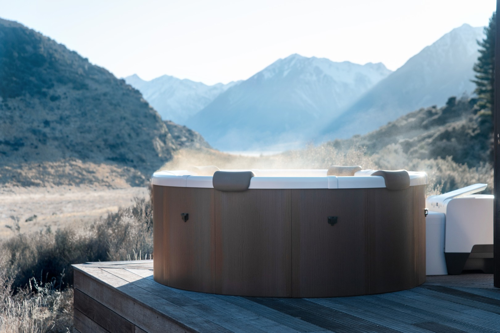 Ein Whirlpool auf einer Holzterrasse mit Bergblick. Dampf steigt aus der Wanne, mit Bergen und Natur im Hintergrund.