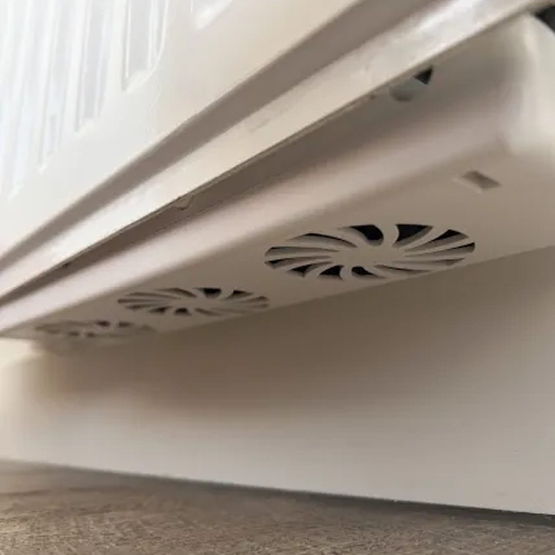 Close-up van een witte radiator met ventilatieopeningen. De radiator is gemonteerd op een witte muur boven een houten vloer.