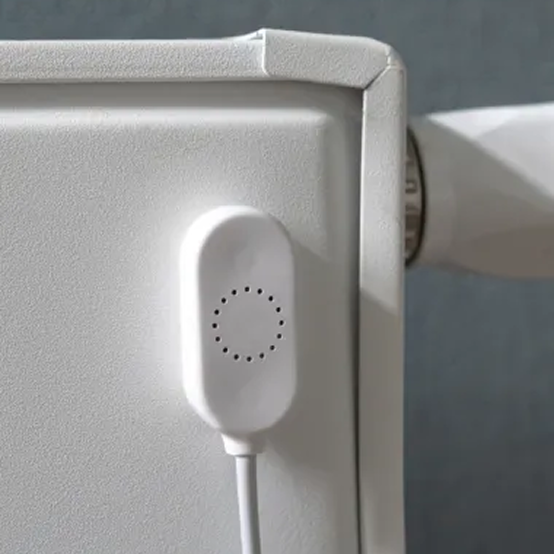 Een wit apparaat met een cirkelvormig patroon van gaten is bevestigd aan een witte radiator. Een witte kabel loopt naar beneden.