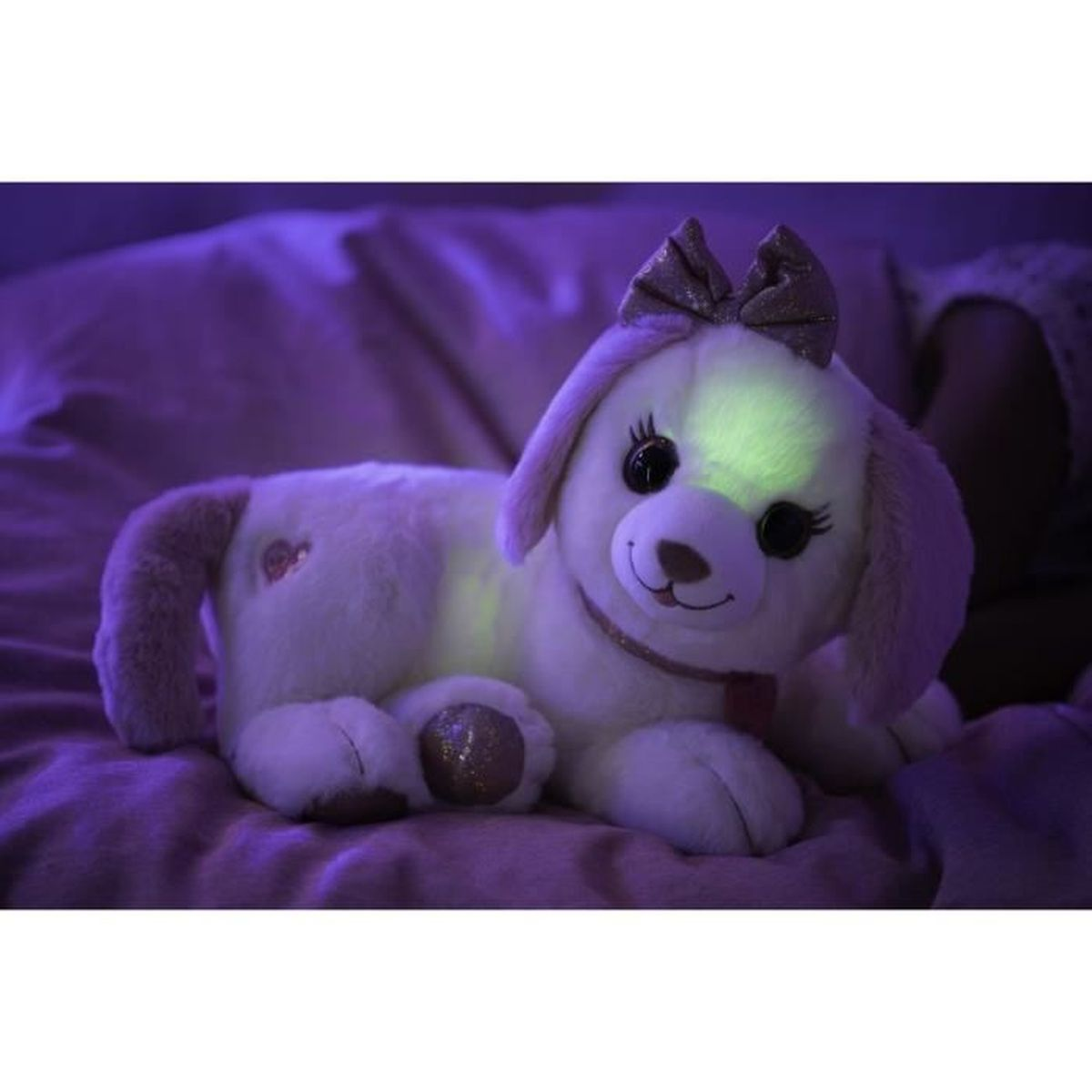 Un jouet en peluche en forme de chien avec un nœud, des éléments lumineux, couché sur une couverture violette. Scène éclairée, personne en arrière-plan.