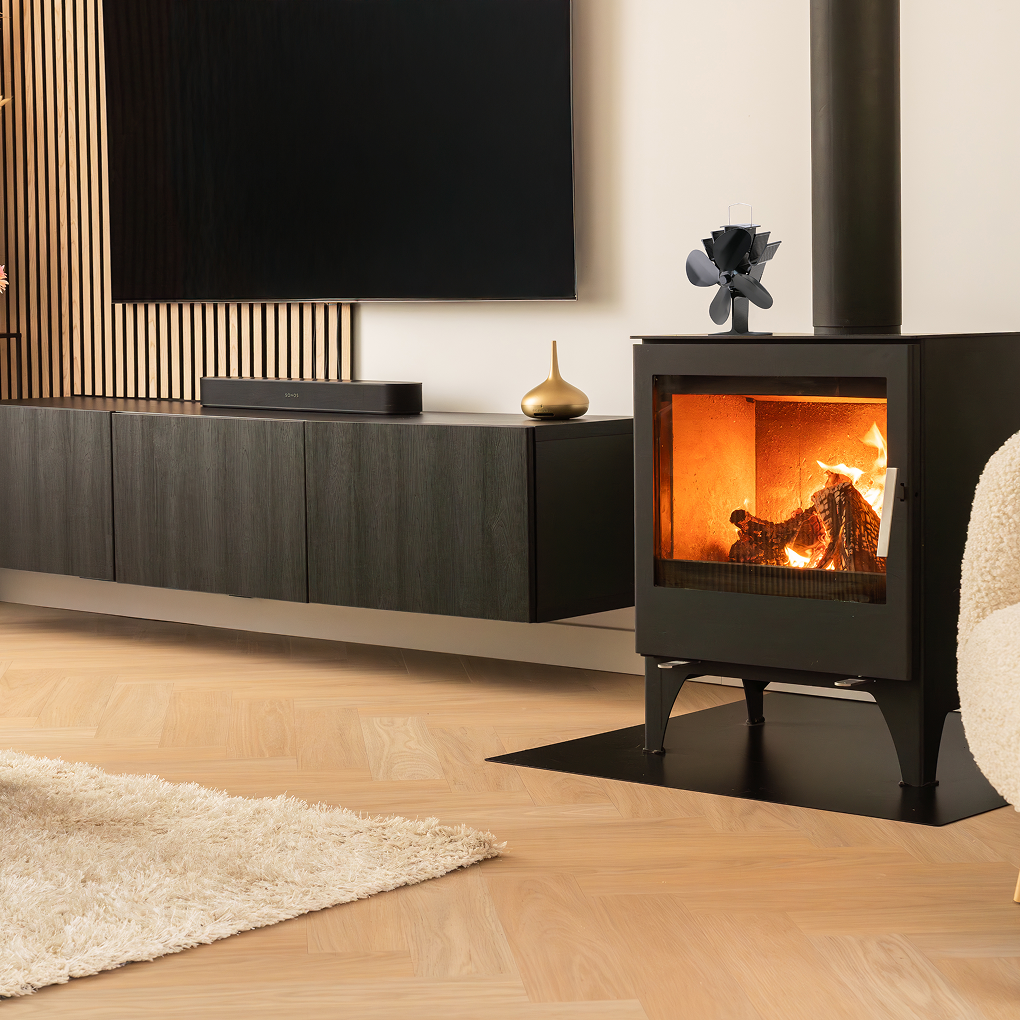 Woonkamer met een brandende open haard, een zwarte tv, een soundbar en een kleed. De vloer is van hout. Er staat een gouden vaas.
