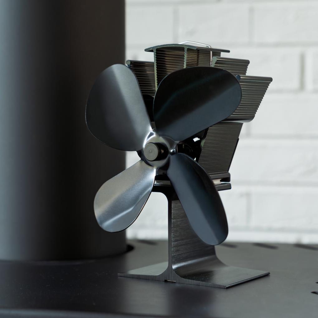 Een zwarte kachelventilator met vier bladen staat in het midden, met een zwarte pijp links en een witte bakstenen muur op de achtergrond.