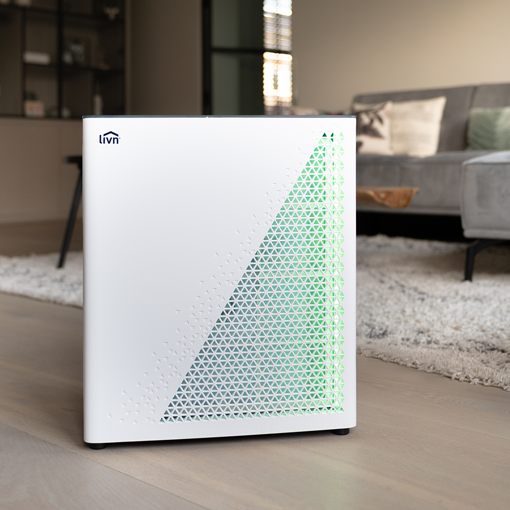 A white air purifier with a green triangle pattern stands in a room. A sofa and rug are in the background.