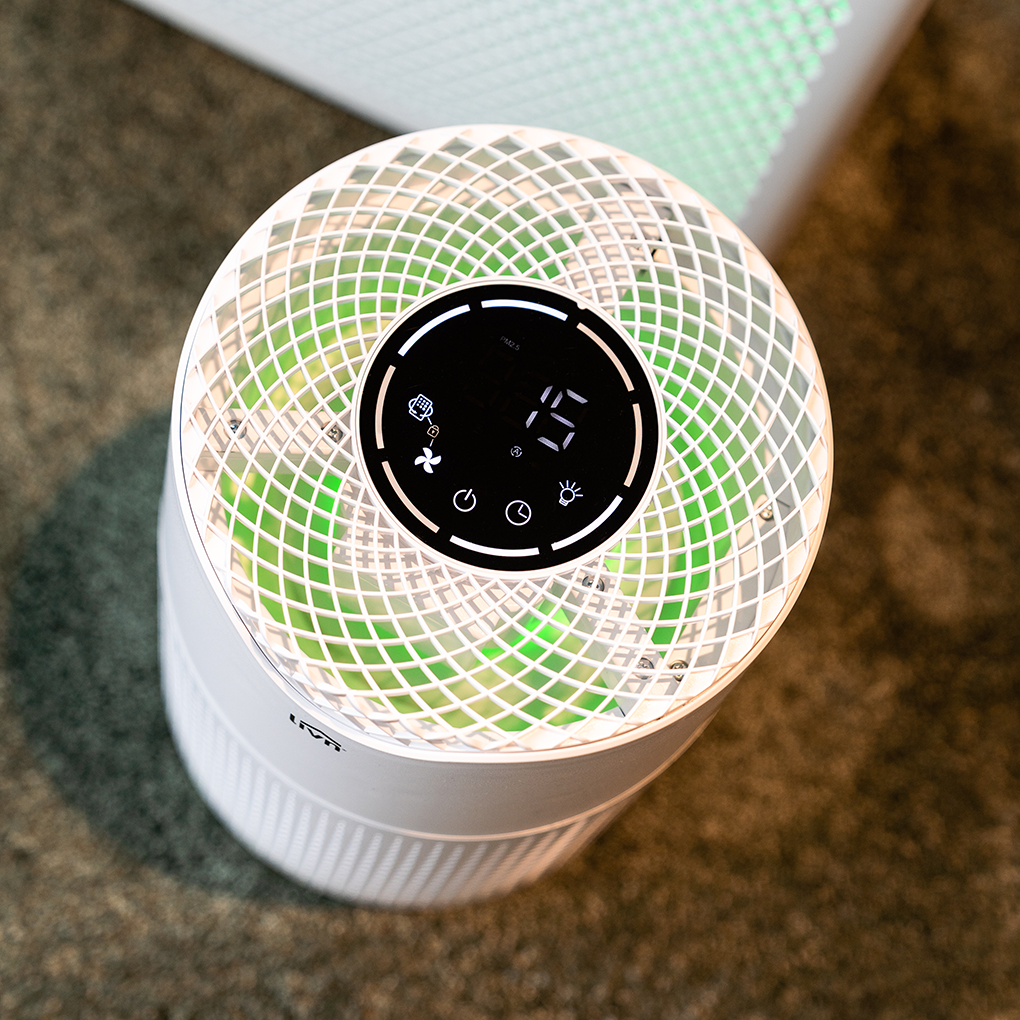 A white air purifier with a digital display showing '15'. It has a grid-like top and green internal lighting. Placed on a carpet.