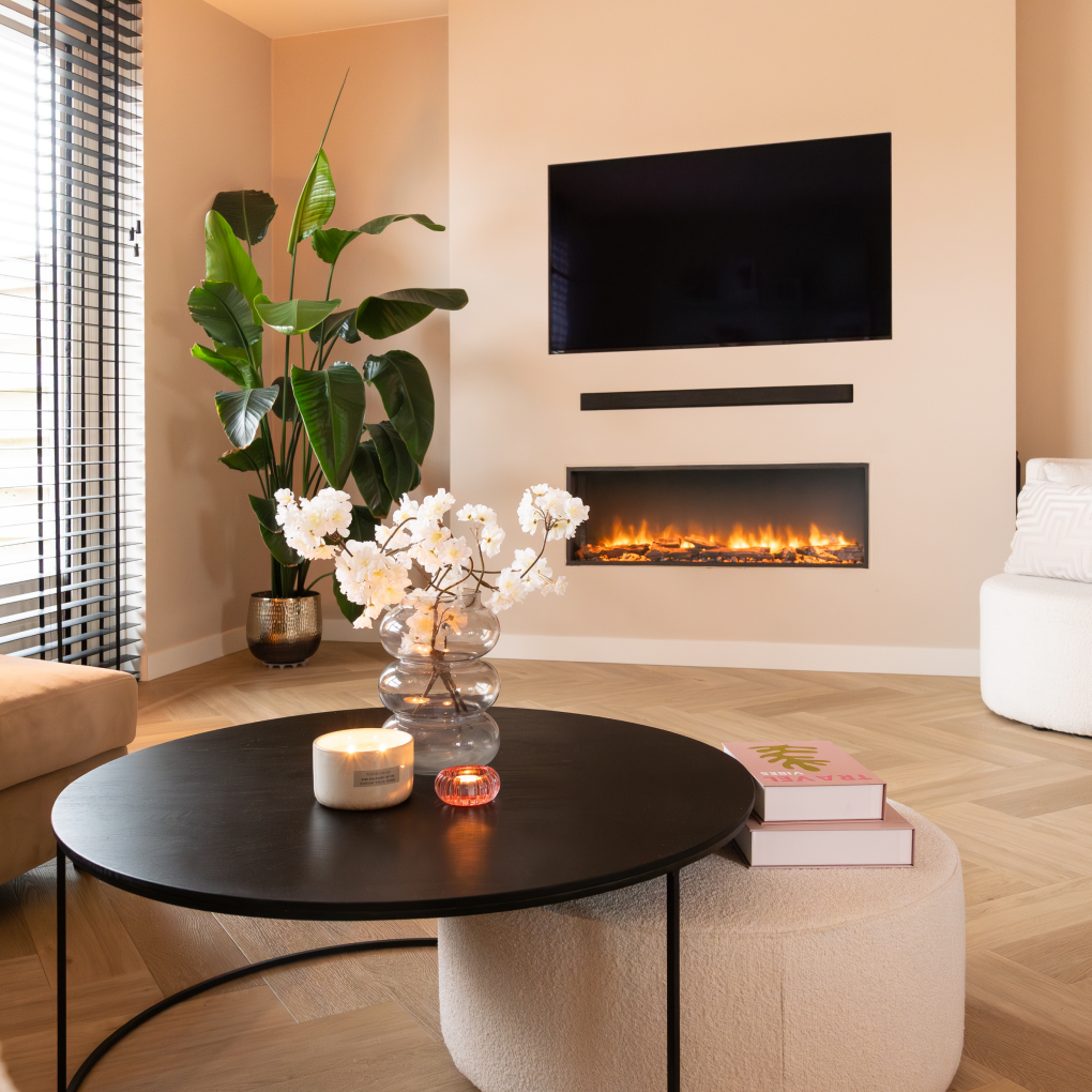 Woonkamer met zwarte tafel, bloemen, open haard, tv en een grote plant in een gouden pot.