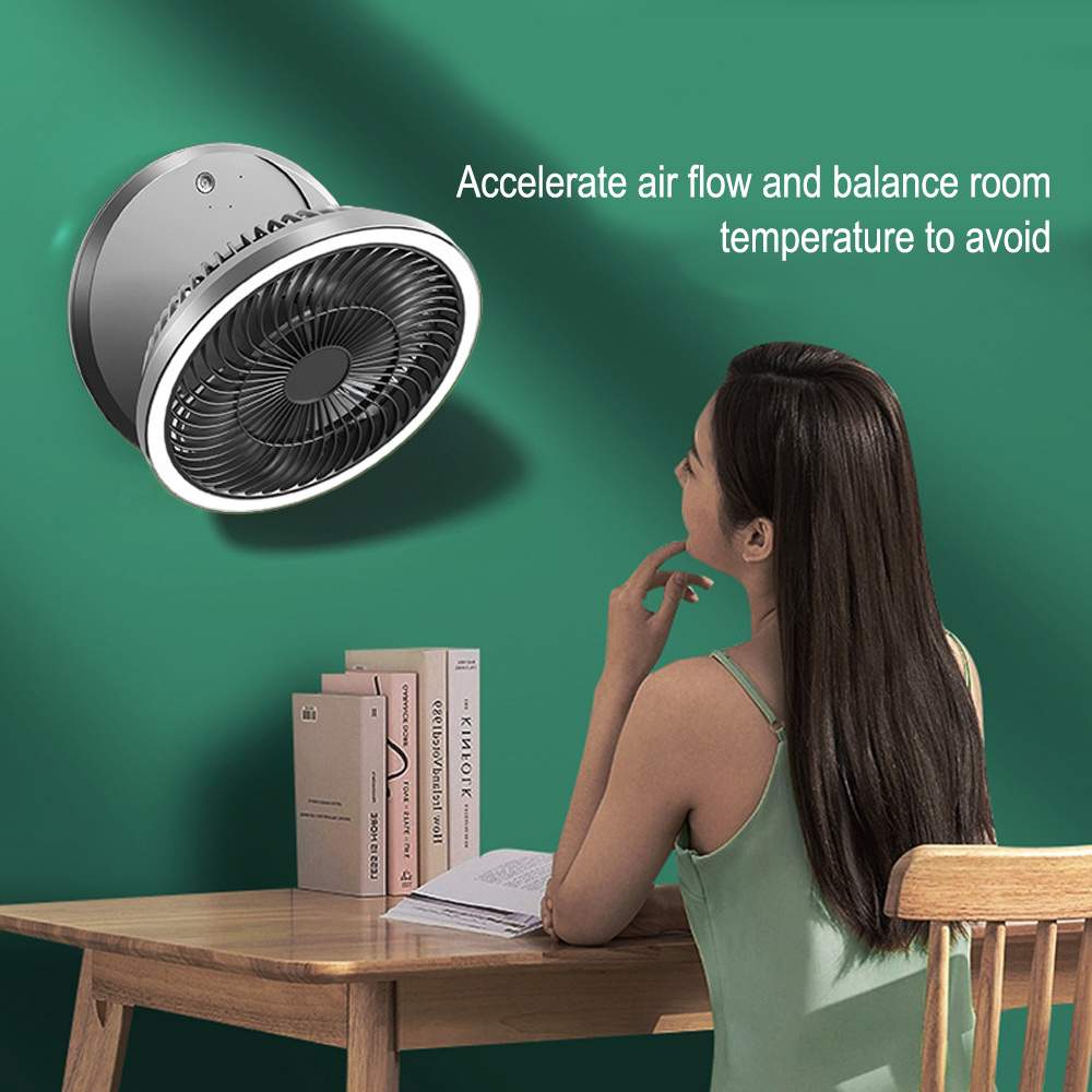 Eine Frau betrachtet einen Ventilator an der Wand. Auf einem Tisch liegen Bücher und Papiere. Der Ventilator ist silber und schwarz, mit weißem Licht.