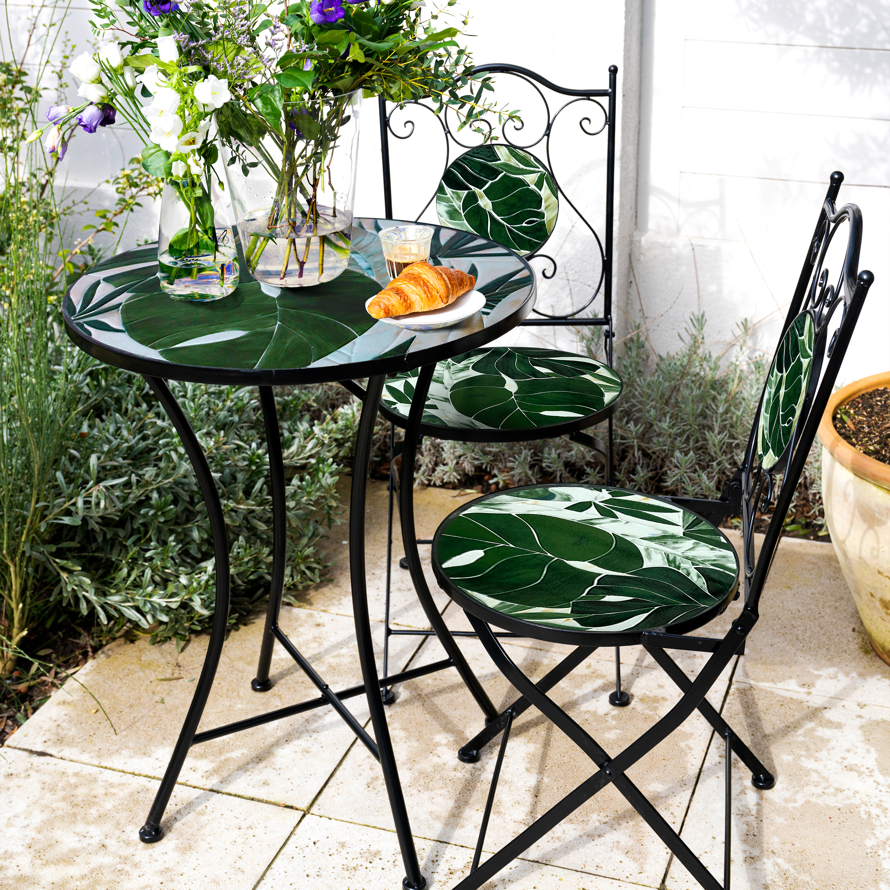 Ein Tisch und zwei Stühle auf einer Terrasse. Der Tisch hat ein florales Design. Ein Croissant liegt auf einem Teller.