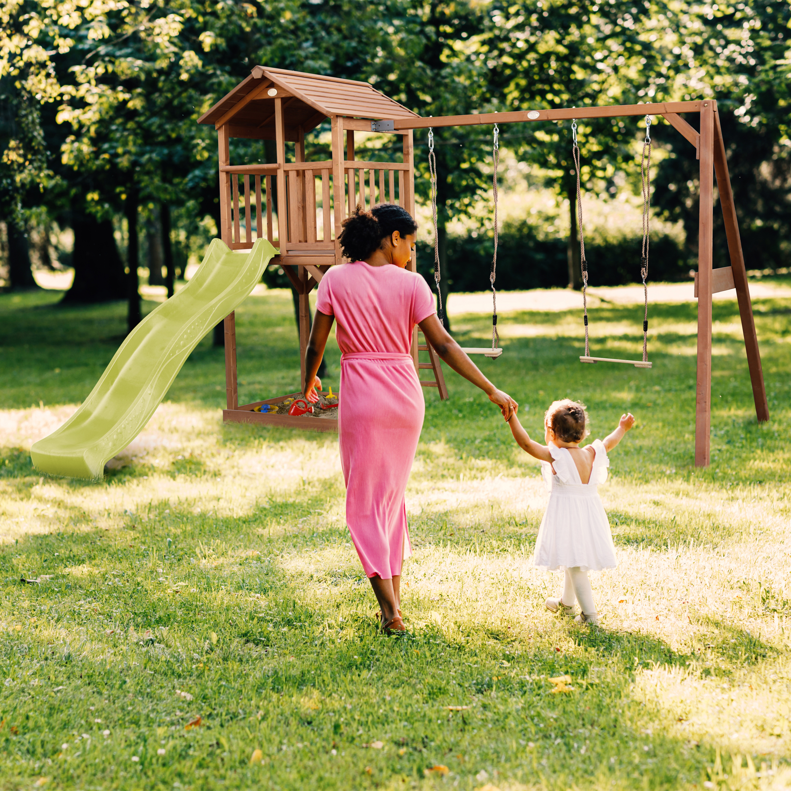 Eine Frau in rosa Kleid hält die Hand eines Kindes in der Nähe eines Spielplatzes mit Rutsche und Schaukeln.