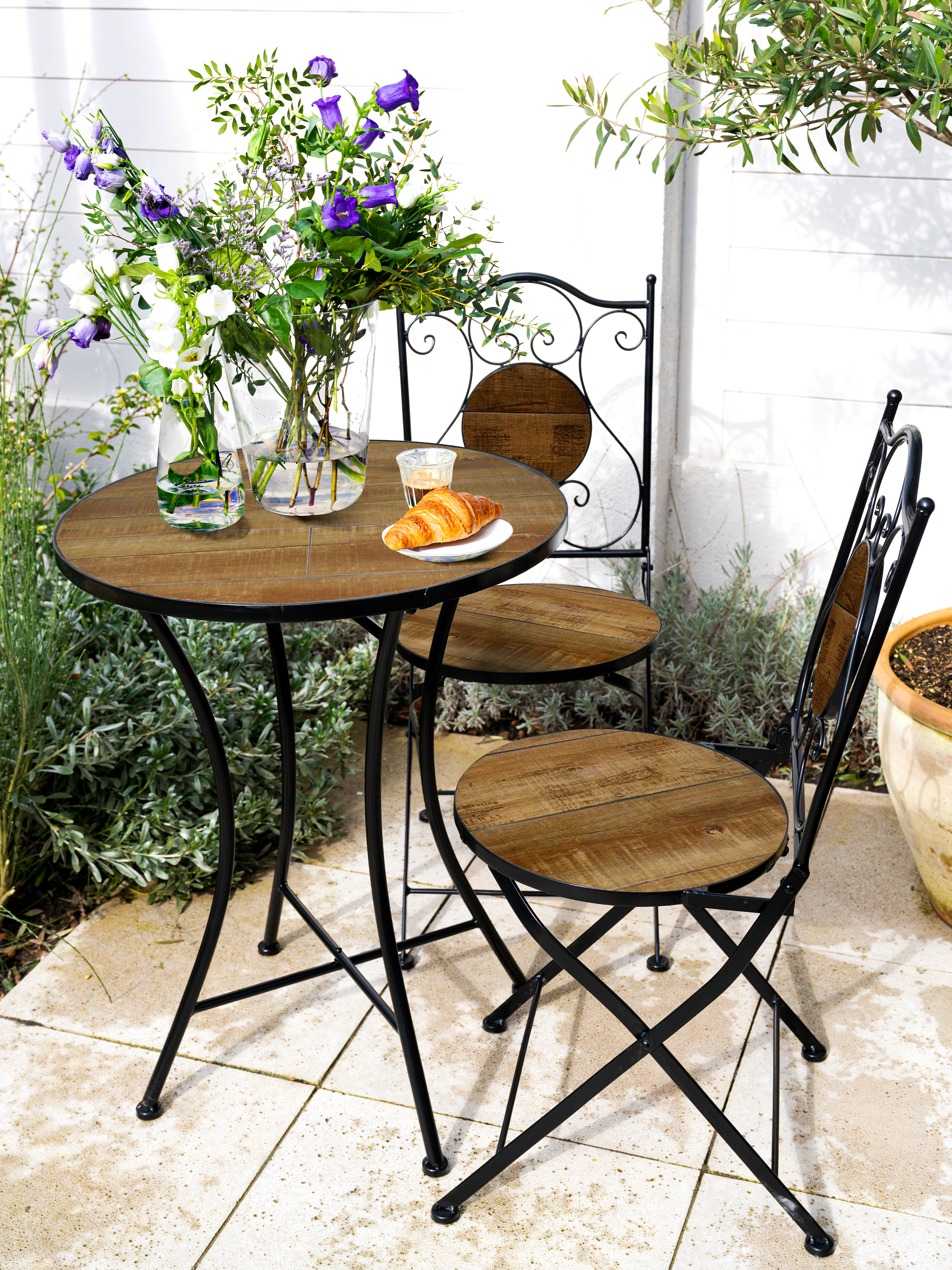 Ein Tisch und zwei Stühle auf einer Terrasse, mit Blumen in einer Vase und einem Croissant auf einem Teller.