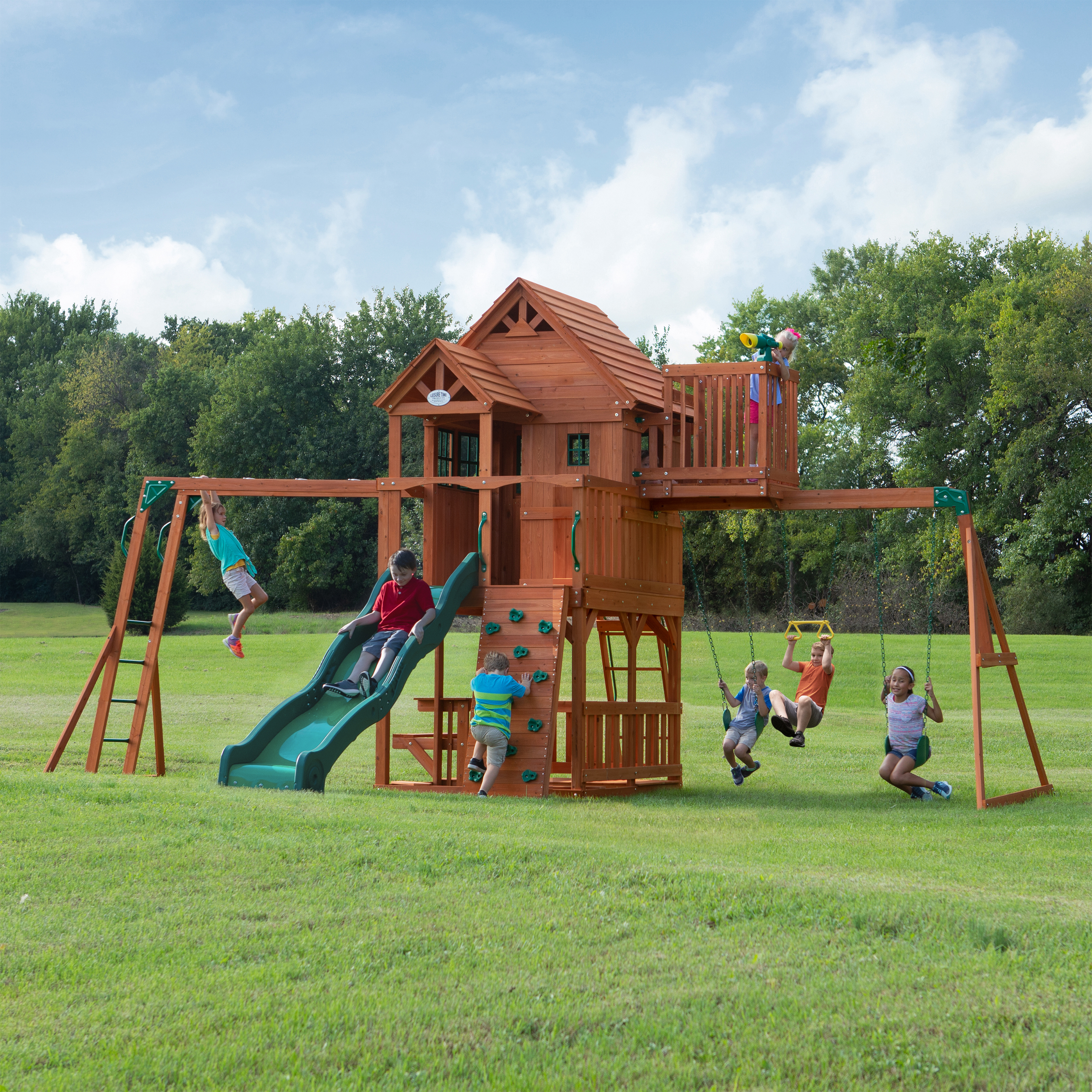 Kinder auf einem Holzspielplatz mit Rutsche, Schaukeln und Kletterwand. Grünes Gras und Bäume im Hintergrund.