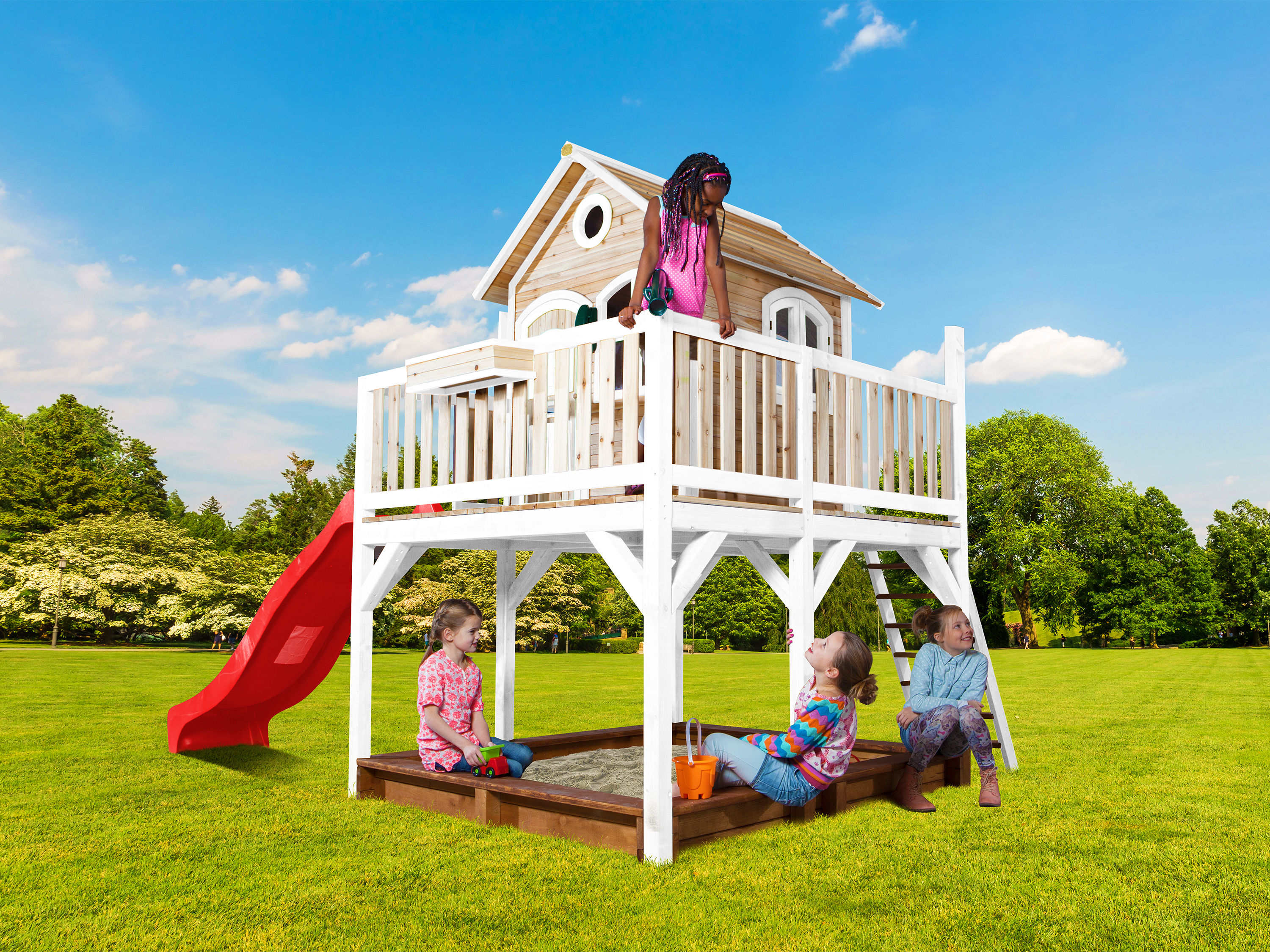 Kinder spielen auf einem Holzspielhaus mit roter Rutsche auf grünem Gras unter blauem Himmel.