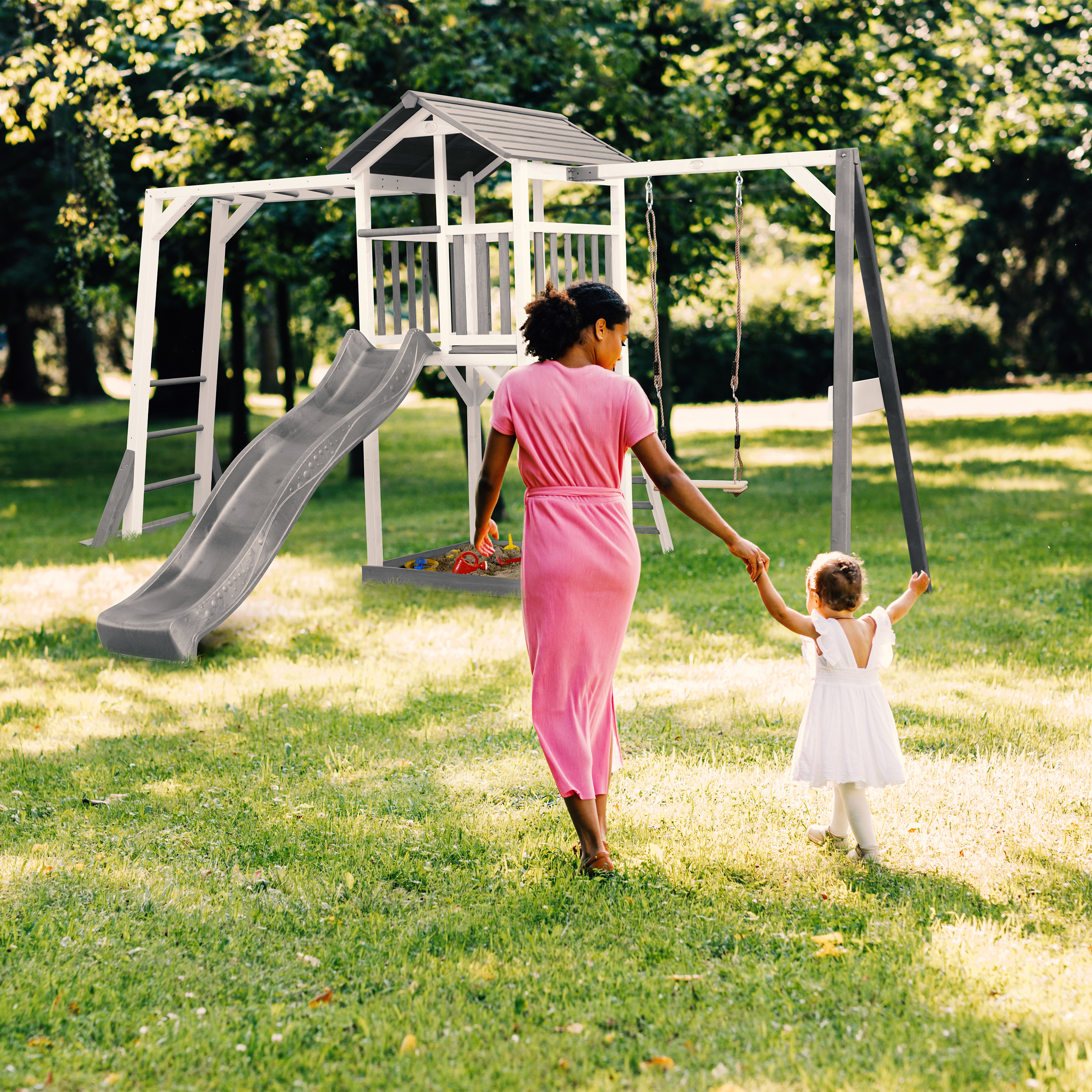 Eine Frau und ein Kind gehen auf einem Spielplatz in einem Park. Die Frau trägt Rosa, das Kind Weiß. Grünes Gras umgibt.