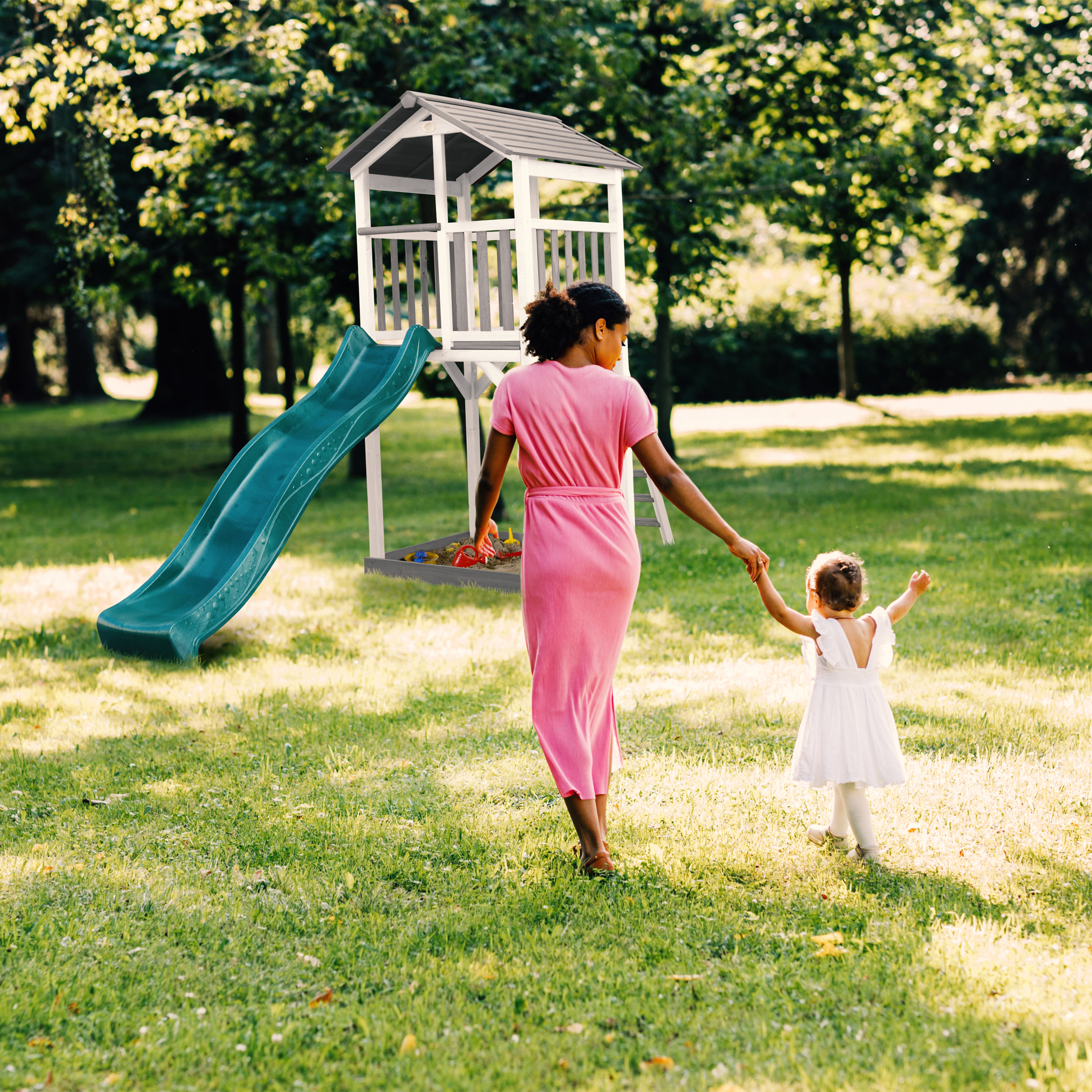 Eine Frau und ein Kleinkind gehen auf Gras zu einem Spielplatz mit einer Rutsche. Bäume sind im Hintergrund.