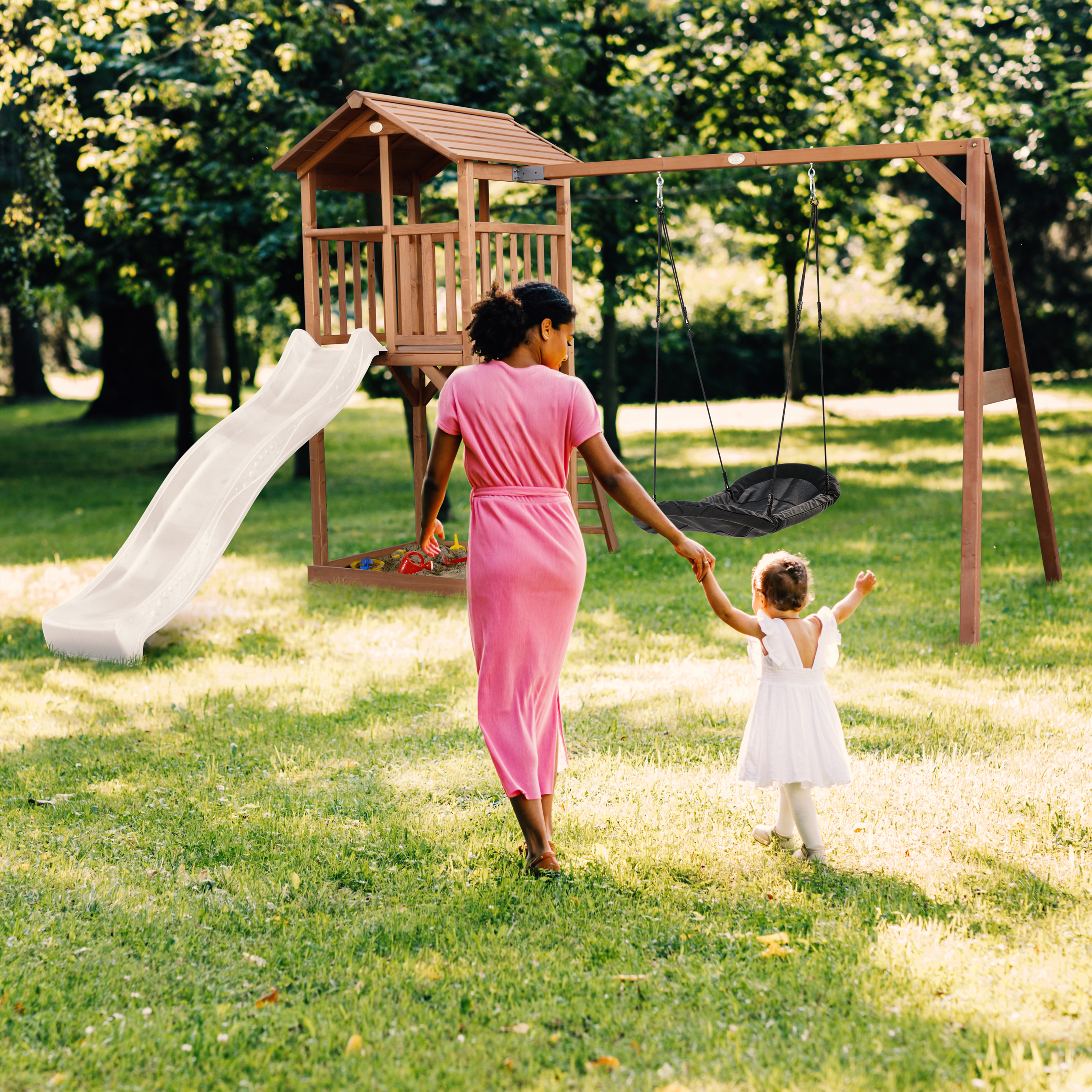 Eine Frau in rosa Kleid hält die Hand eines Kindes und geht zu einem Spielplatz. Eine Schaukel und Rutsche sind im Hintergrund.