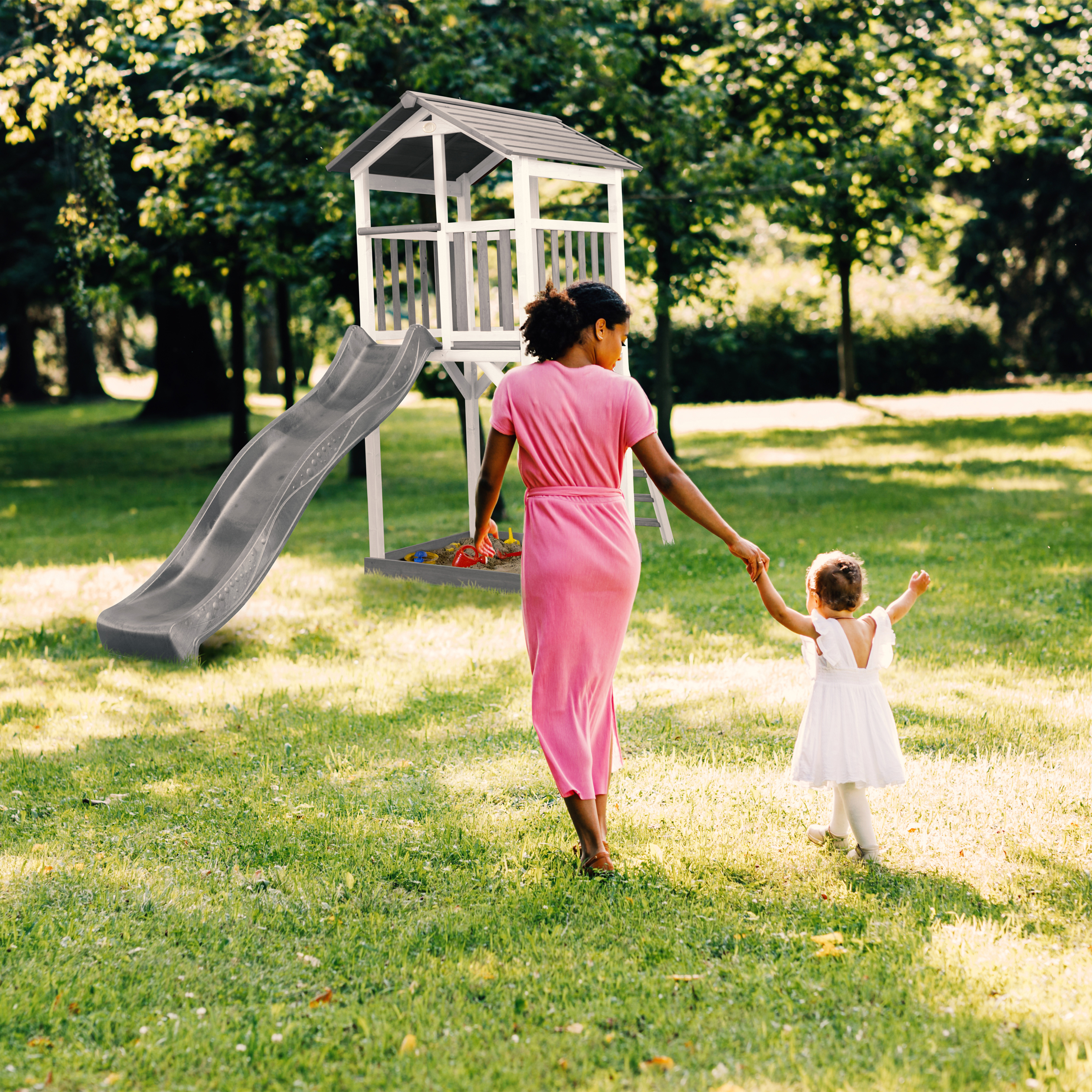 Eine Frau in rosa Kleid und ein Kind in weißem Kleid gehen auf einen Spielplatz zu. Grünes Gras und Bäume sind im Hintergrund.
