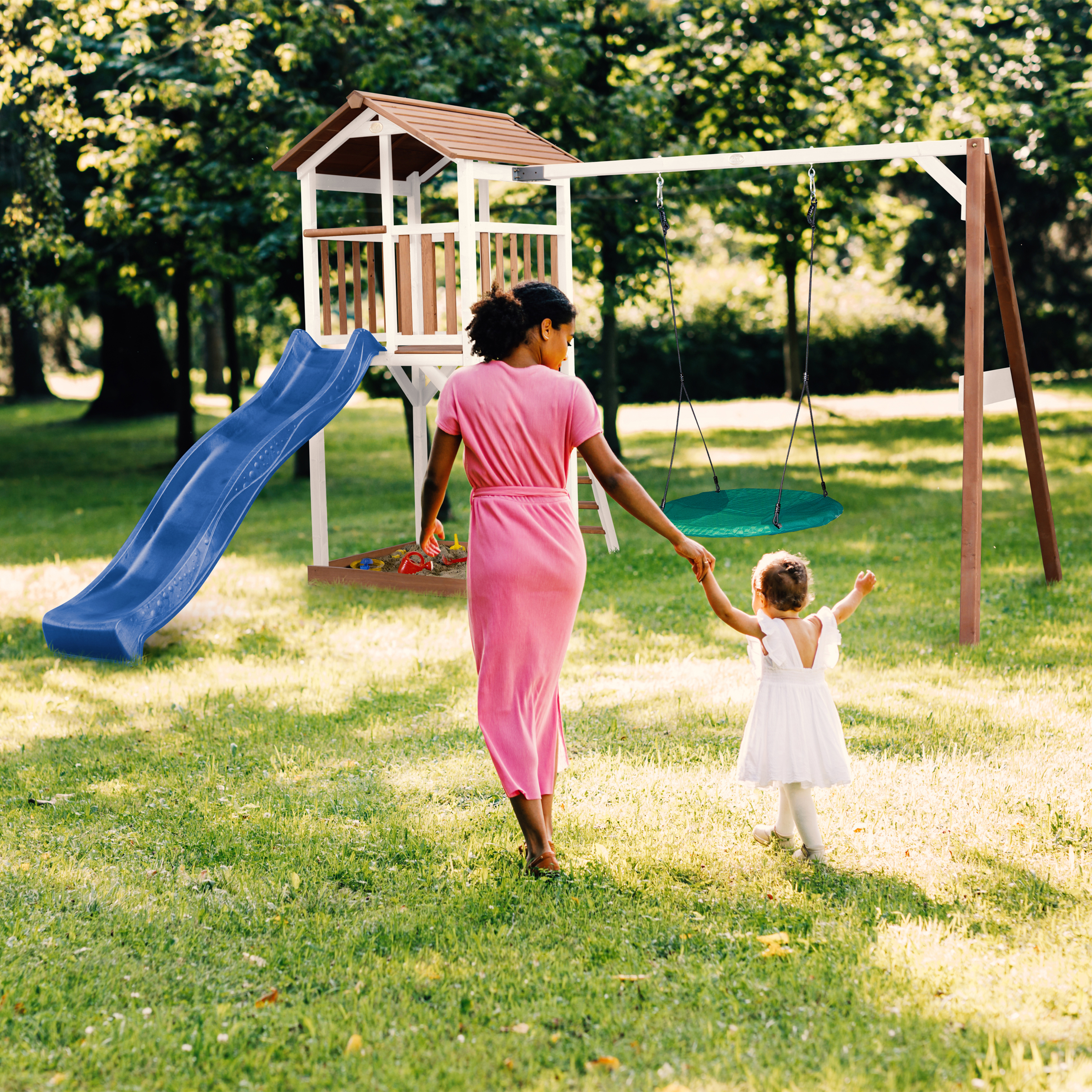 Eine Frau und ein Kind gehen auf einen Spielplatz mit Rutsche und Schaukel auf einer grünen Wiese zu.