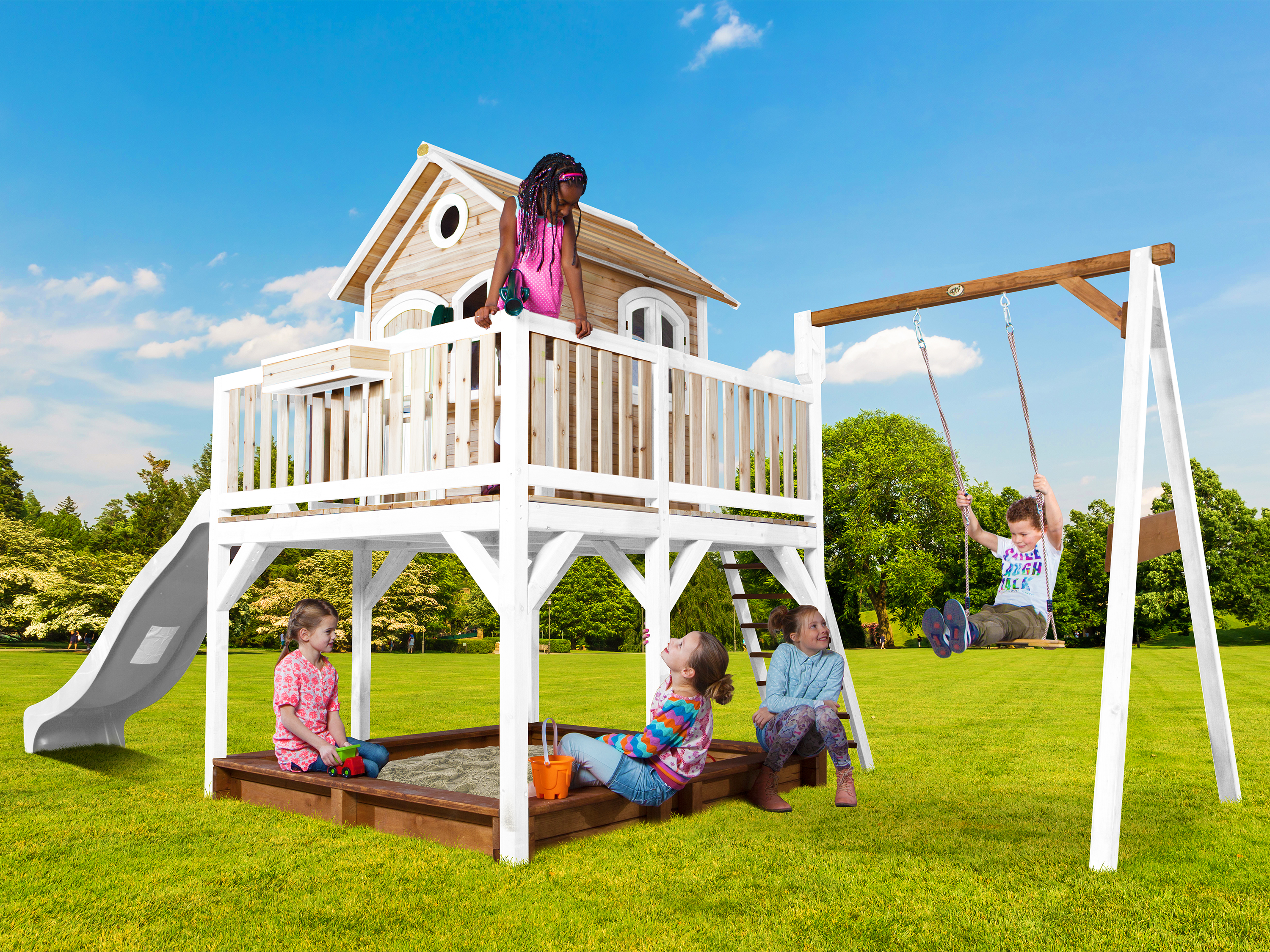 Kinder spielen auf einem Spielplatz mit einem Spielhaus, einer Schaukel und einem Sandkasten. Blauer Himmel und grünes Gras im Hintergrund.