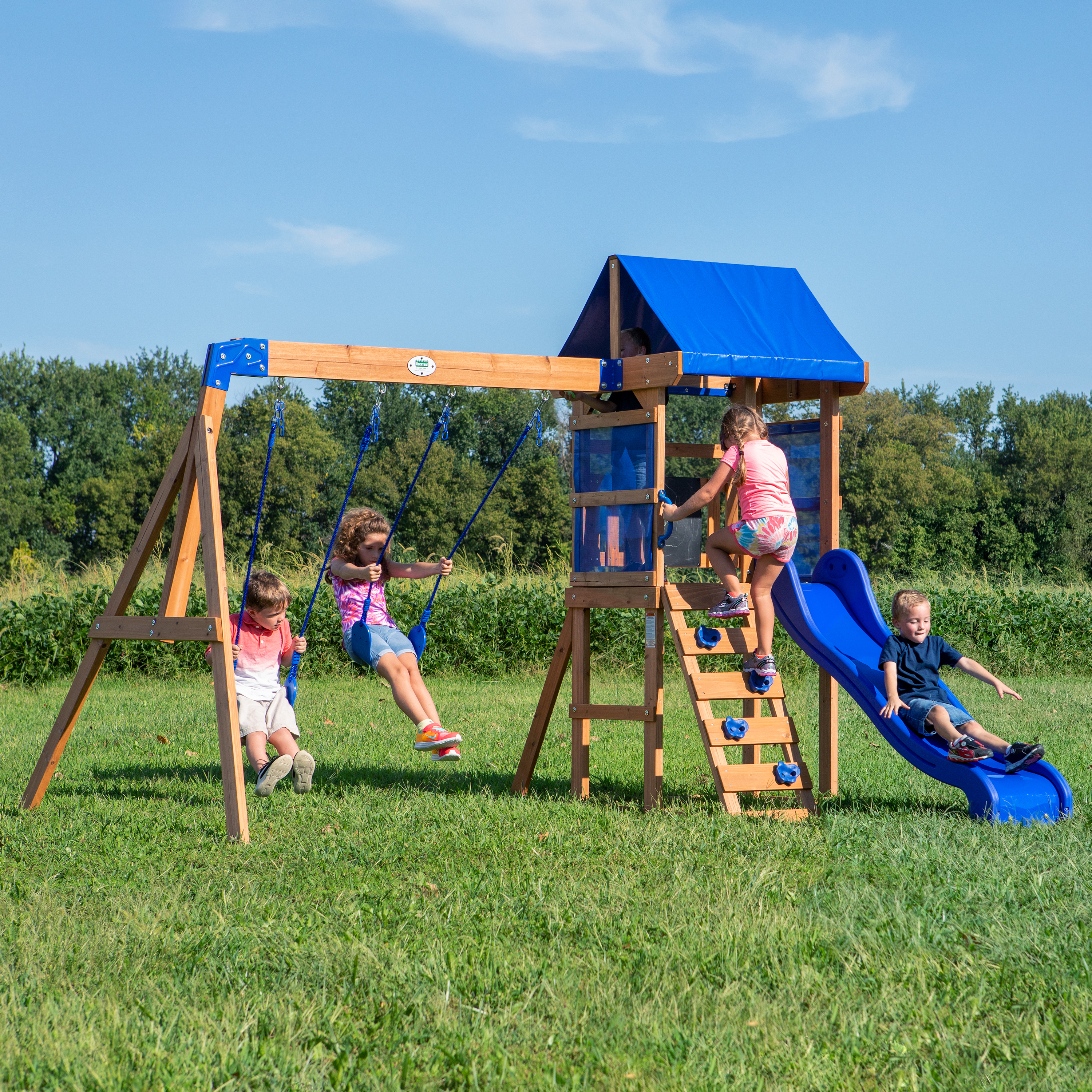 Kinder spielen auf einem Holzspielplatz mit Schaukeln, Rutsche und Kletterwand. Blauer Himmel und grünes Gras.