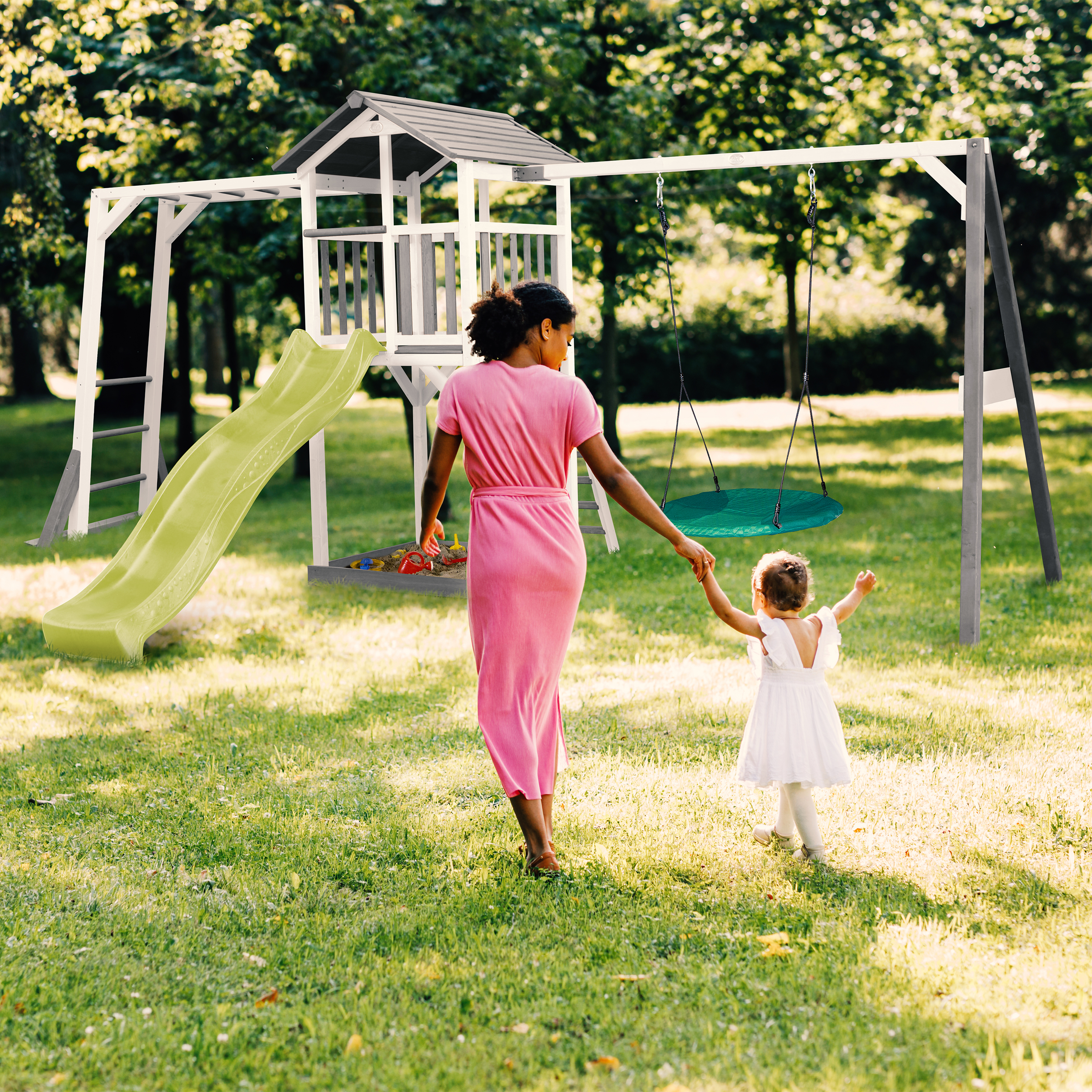 Eine Frau und ein Kind gehen auf einen Spielplatz zu. Der Spielplatz hat eine Rutsche und Schaukeln. Grünes Gras und Bäume umgeben sie.