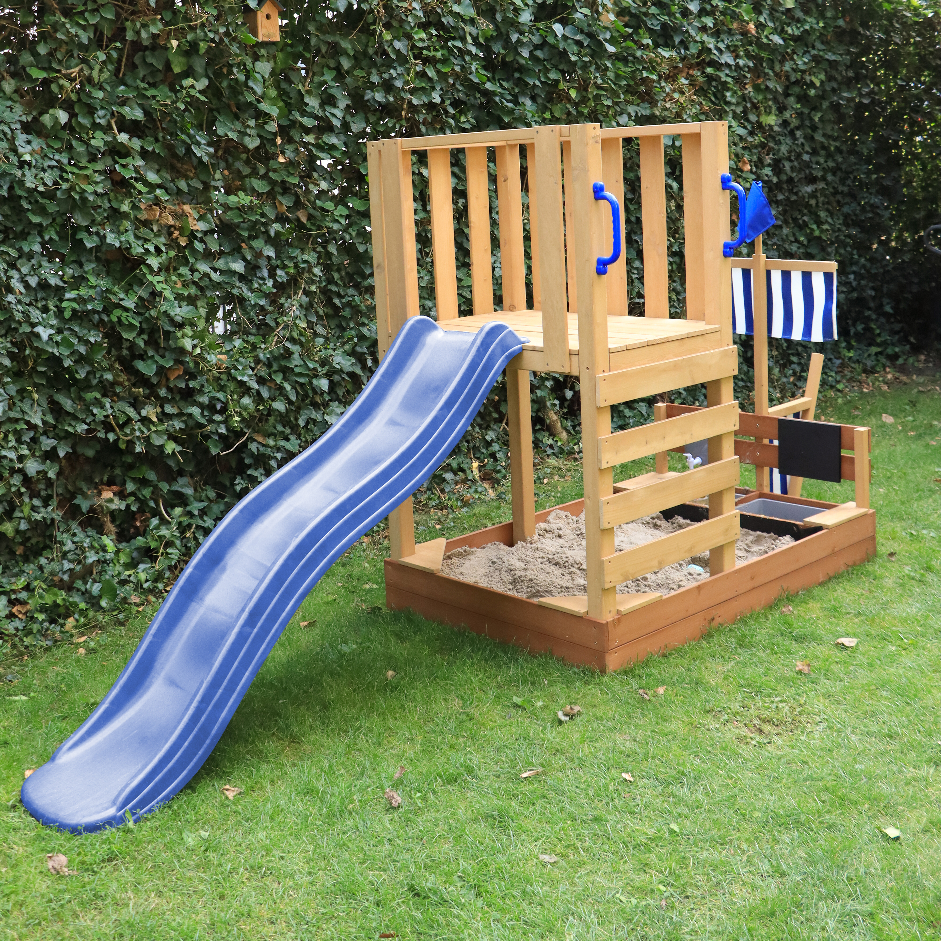 Ein hölzernes Spielset mit blauer Rutsche, Sandkasten und Flagge auf grünem Gras mit Laub im Hintergrund.