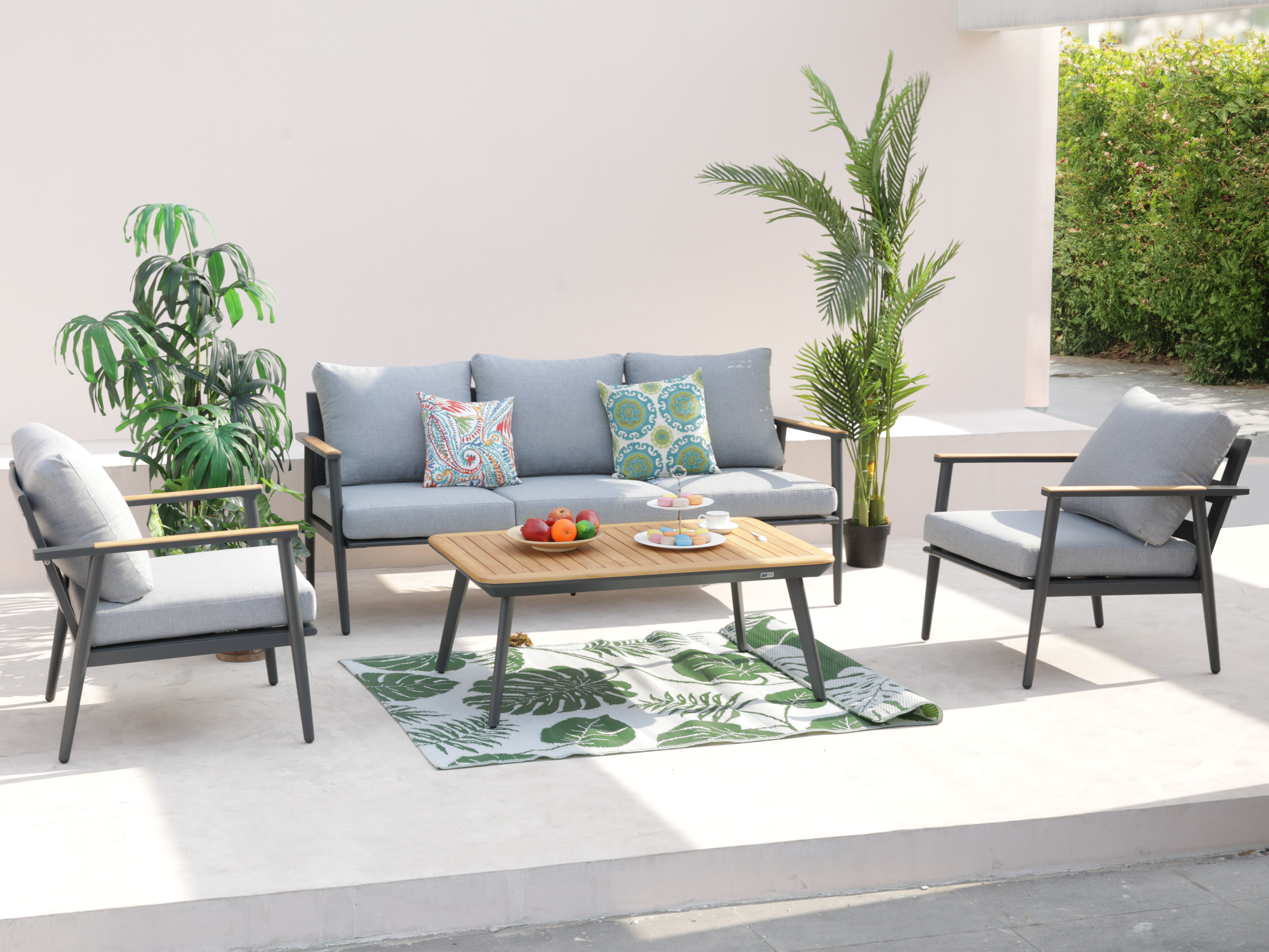Outdoor-Sitzgruppe mit grauen Kissen, Holztisch und tropischem Teppich. Pflanzen umrahmen die Terrasse.