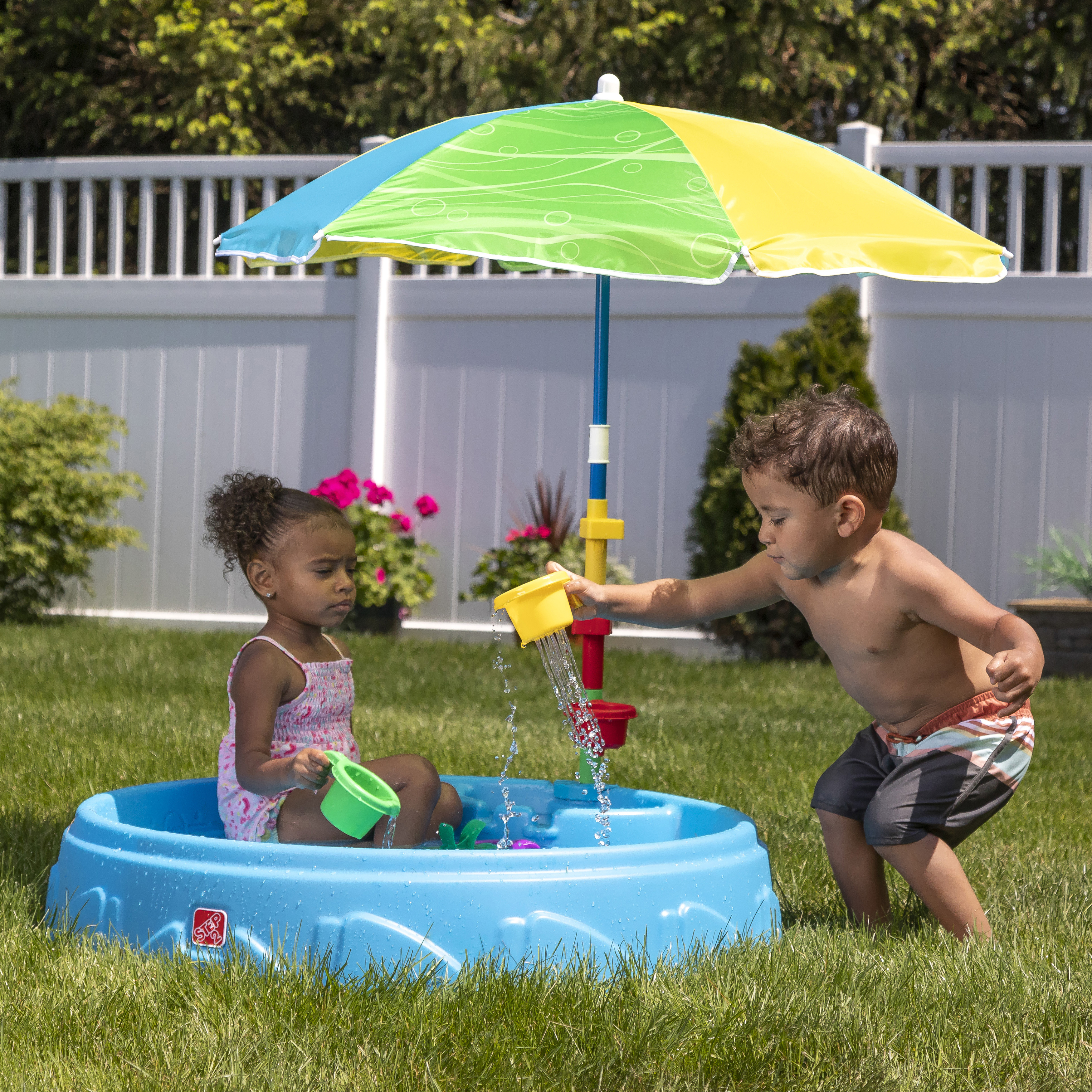 Zwei Kinder spielen in einem blauen Pool unter einem bunten Schirm. Einer gießt Wasser, der andere sitzt.