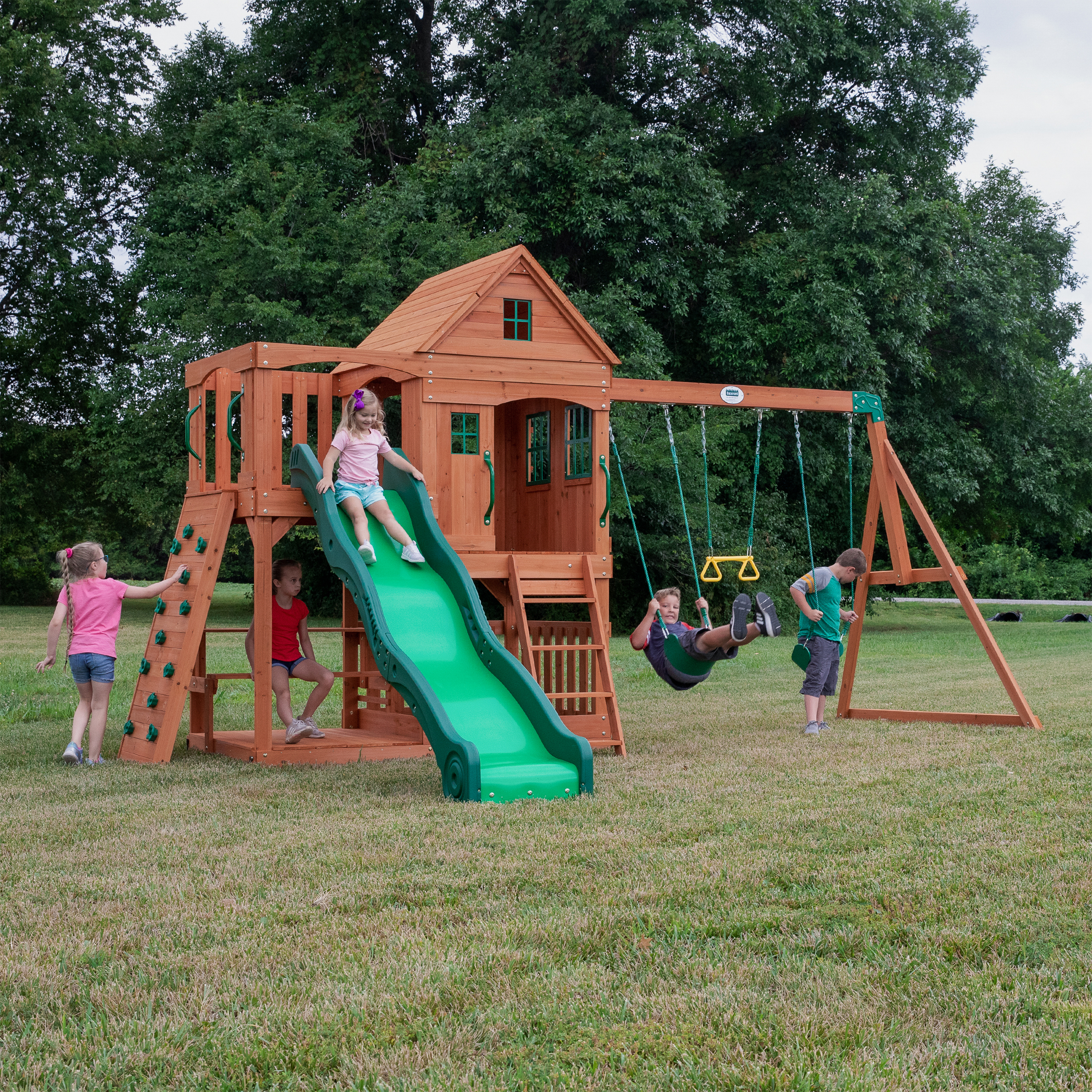 Kinder spielen auf einem Holzspielplatz mit Rutsche, Schaukeln und Kletterwand in einem grasbewachsenen Garten.
