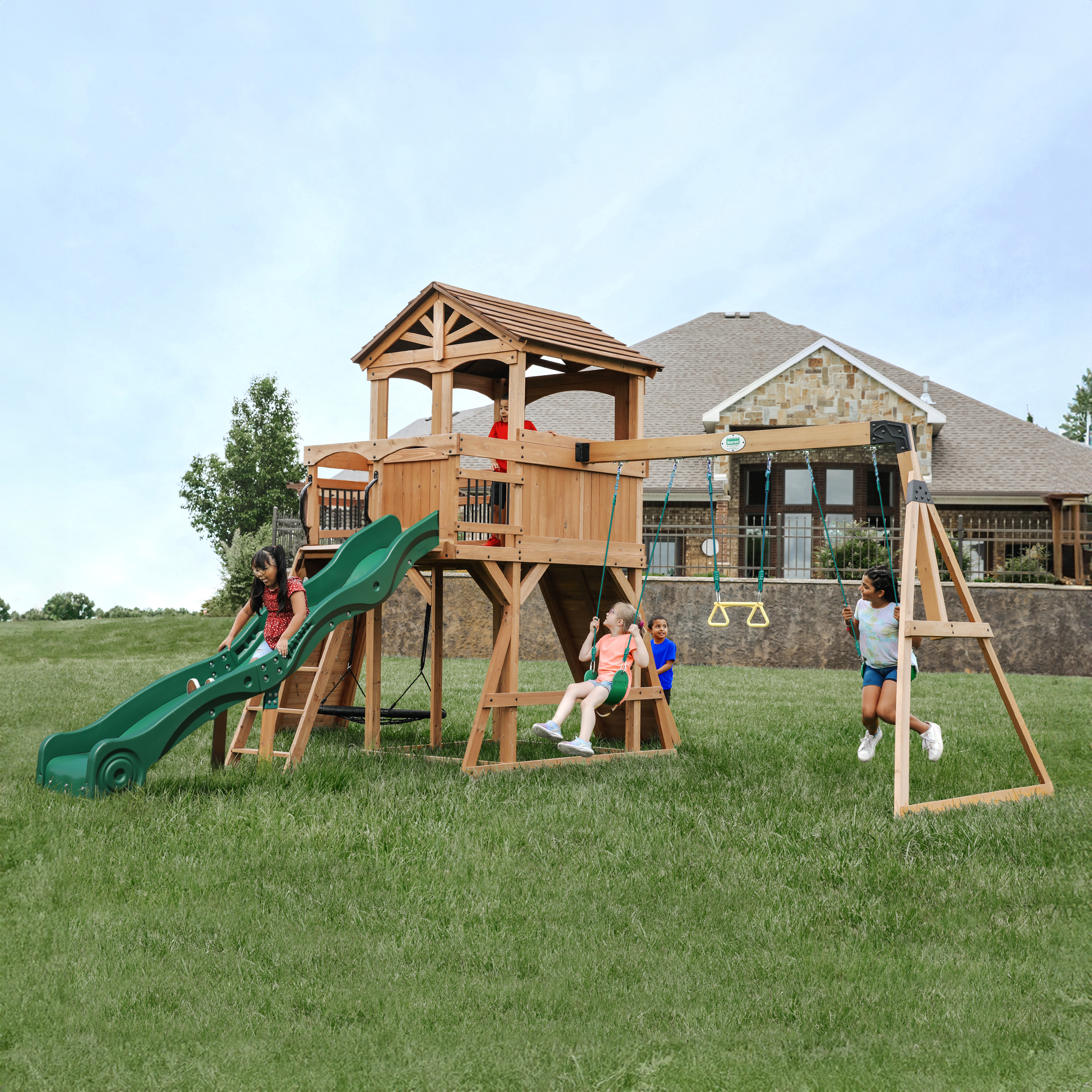 Kinder spielen auf einem Holzspielplatz mit Rutsche und Schaukeln, auf grünem Gras, ein Haus im Hintergrund.