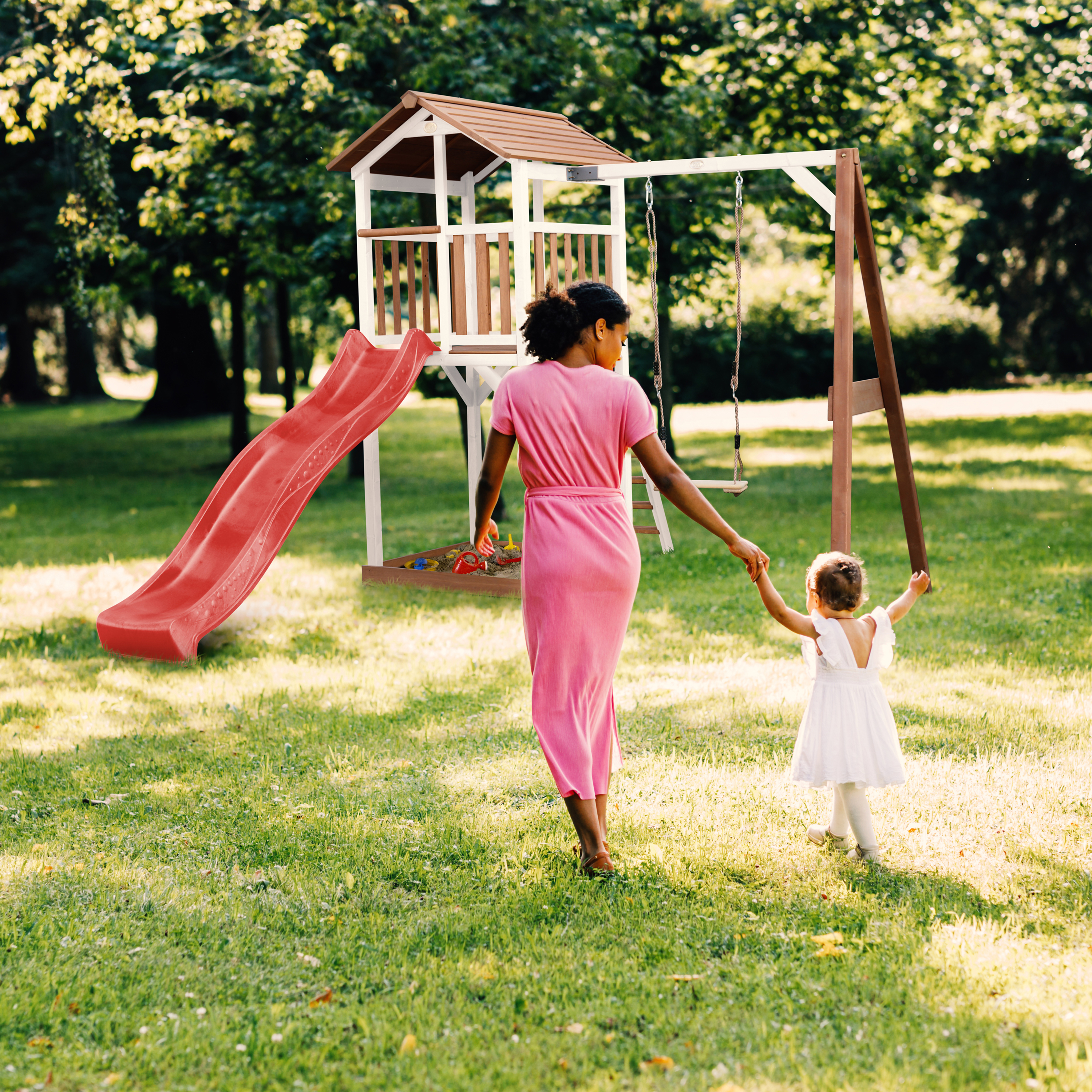 Eine Frau und ein Kind gehen auf einer grünen Wiese auf einen Spielplatz zu. Der Spielplatz hat eine Rutsche und Schaukeln.