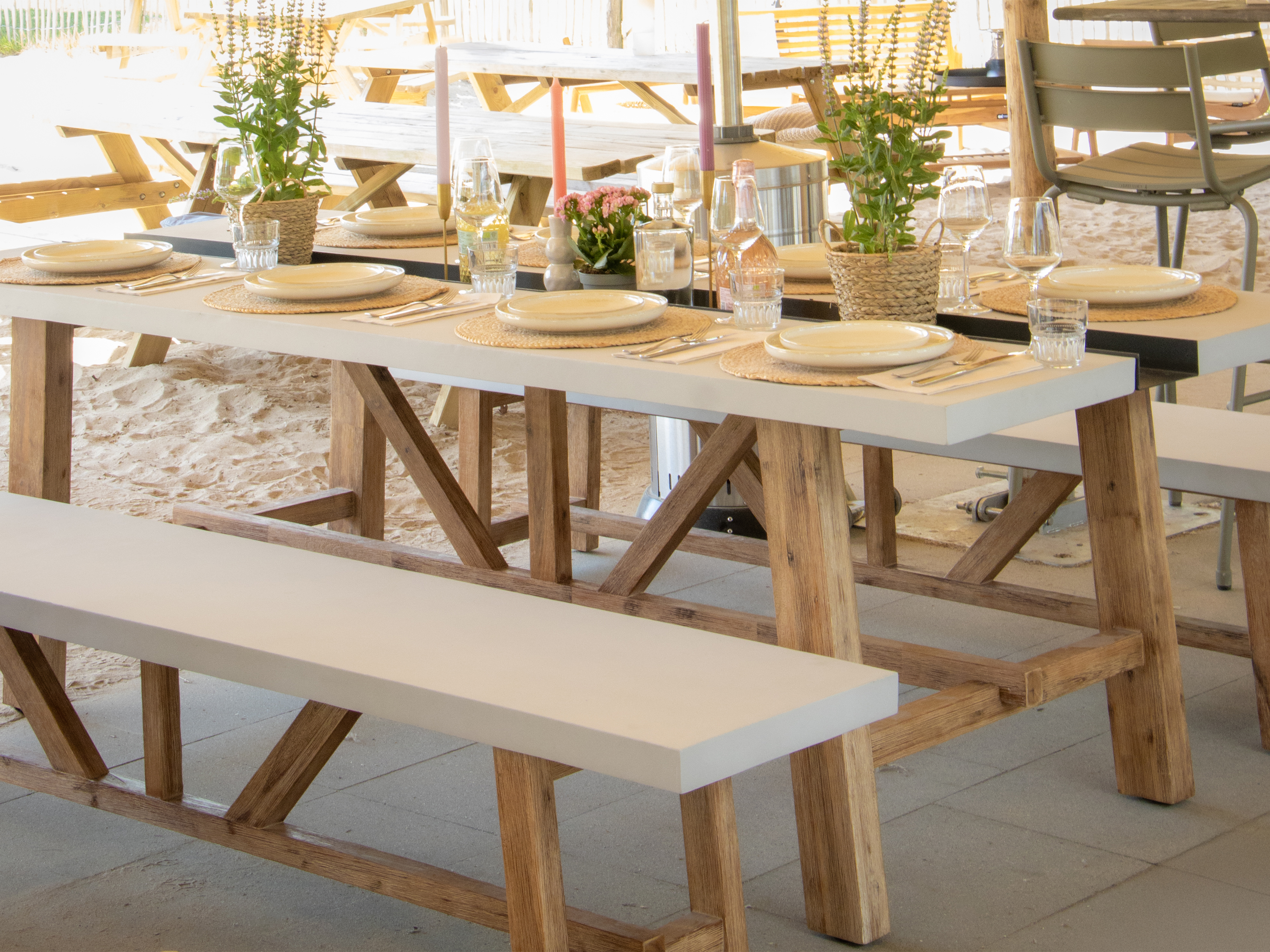Esszimmertisch im Freien. Weiße Tische mit Holzbeinen, gedeckt. Pflanzen und Gläser stehen auf dem Tisch.