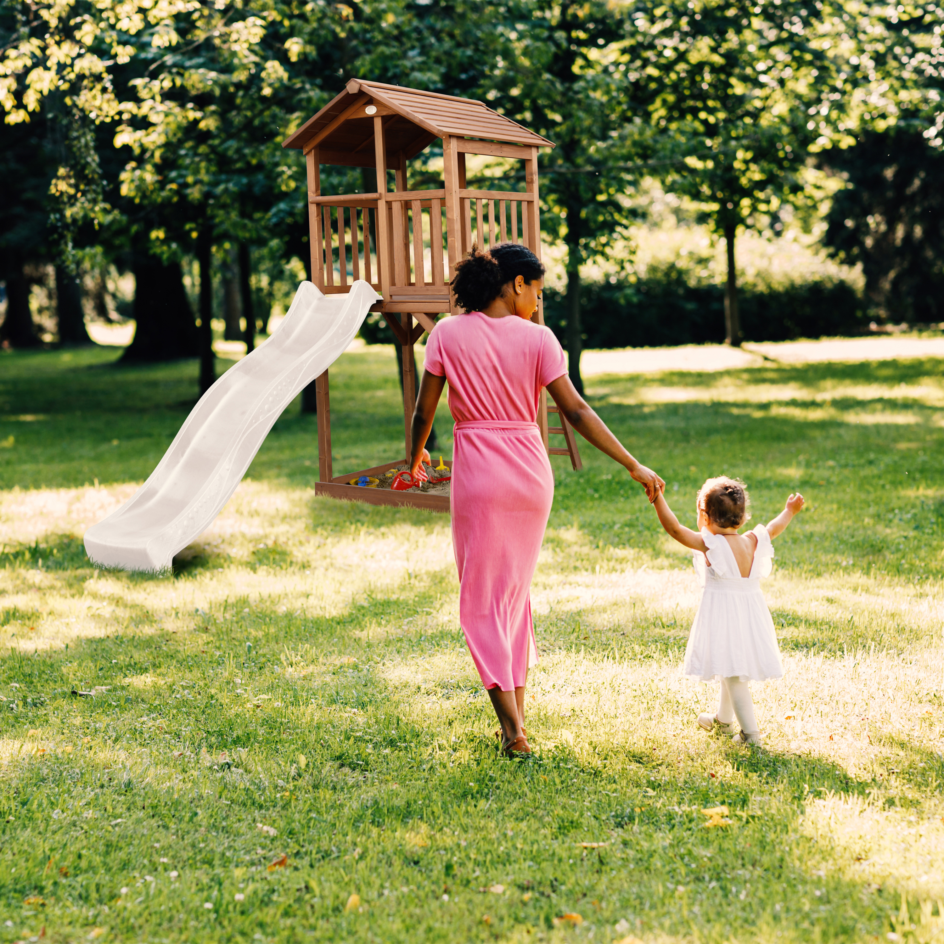 Eine Frau in rosa Kleid hält die Hand eines Kindes und geht in einem Park in der Nähe eines Spielhauses mit Rutsche.