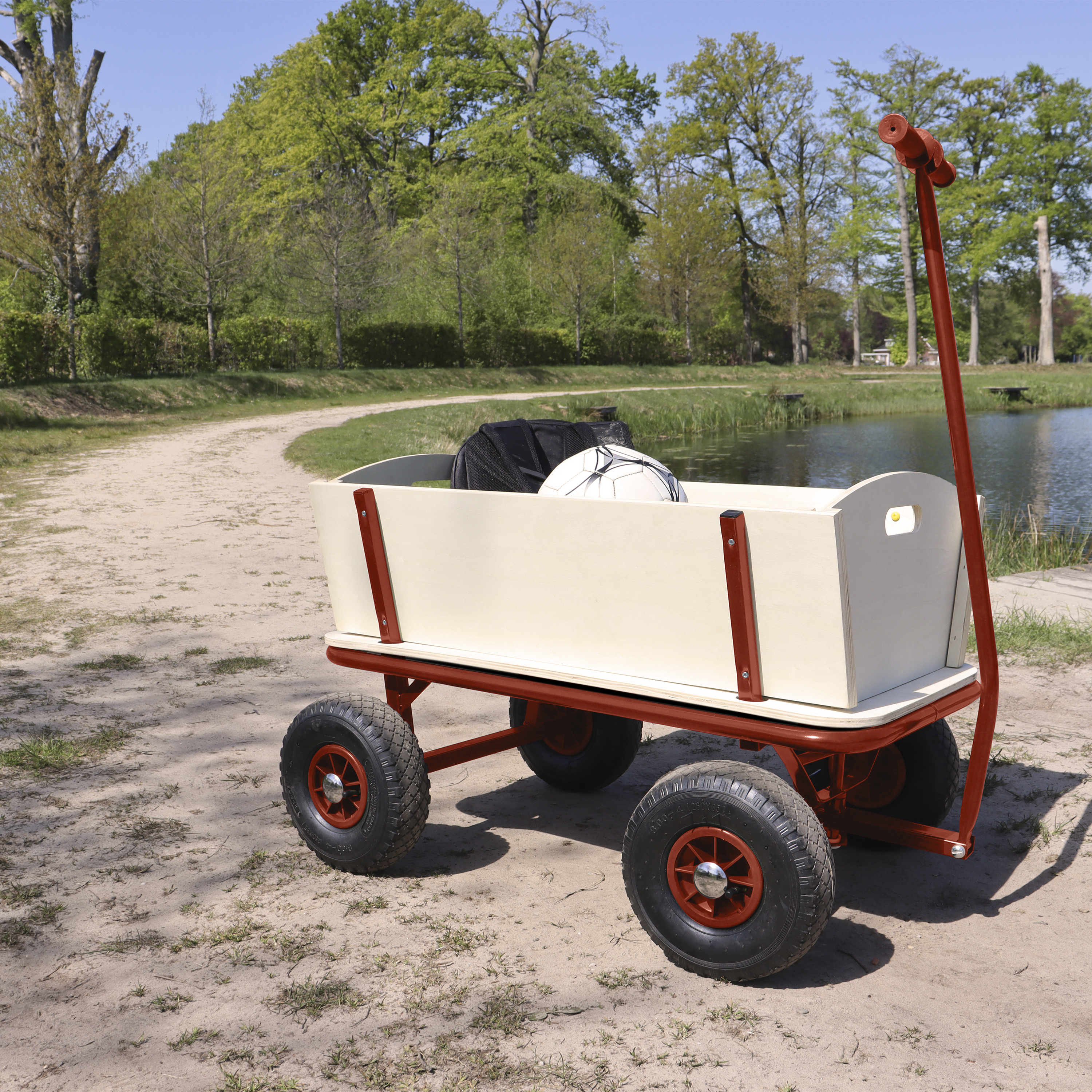 Roter Wagen mit Fußball und Rucksack steht auf Sand neben Wasser und Bäumen. Blauer Himmel im Hintergrund.