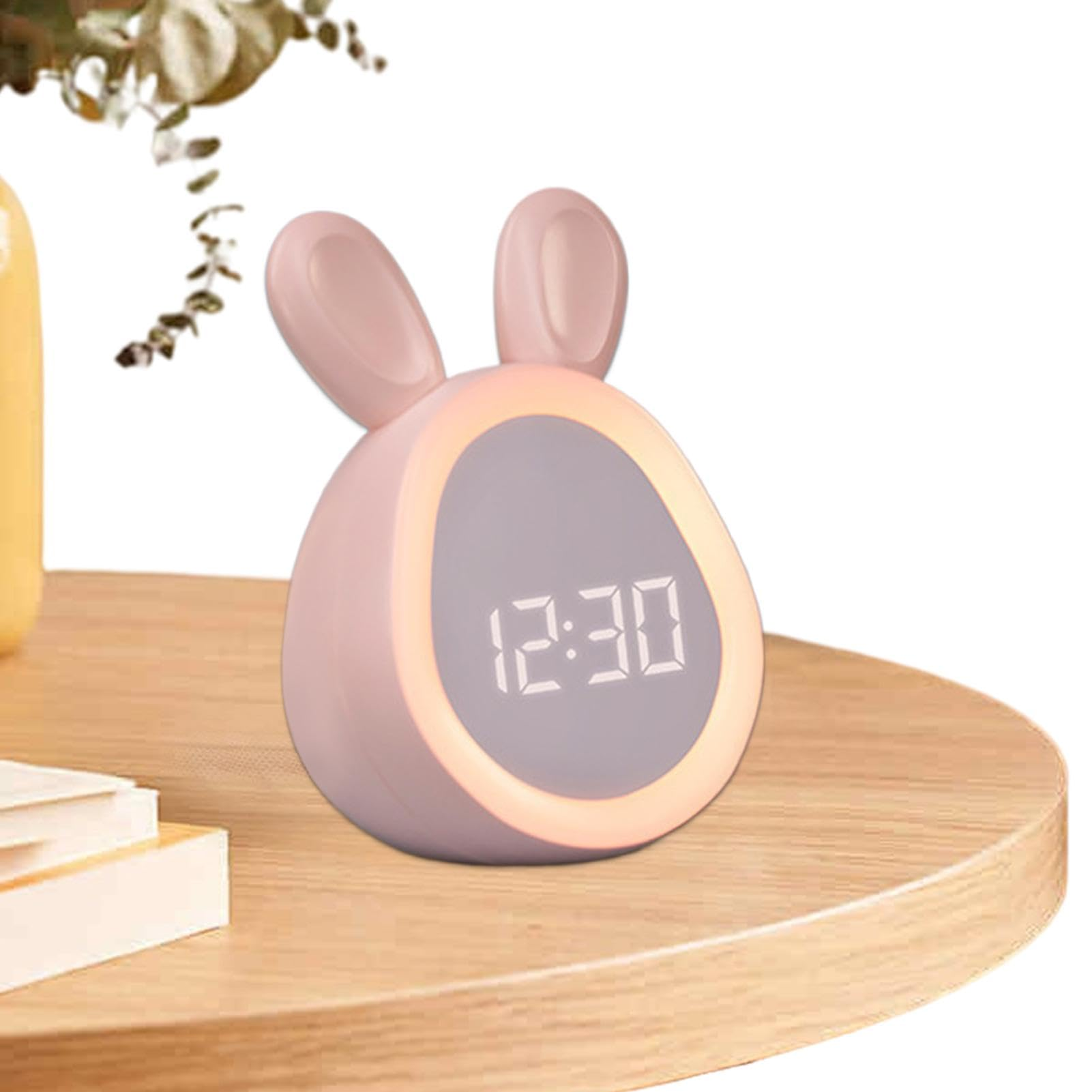 Ein rosa, hasenförmiger Wecker zeigt 12:30 Uhr an. Er steht auf einer Holzoberfläche, im Hintergrund eine gelbe Vase und Pflanze.