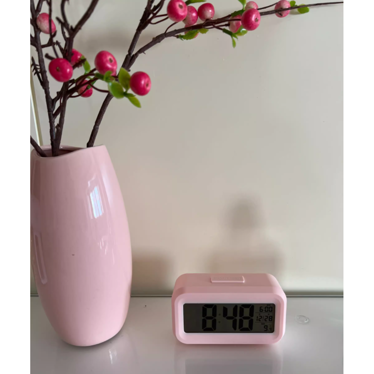 Vaso rosa con rami e bacche rosse. Un orologio digitale rosa mostra le 8:48. Lo sfondo è una parete bianca.