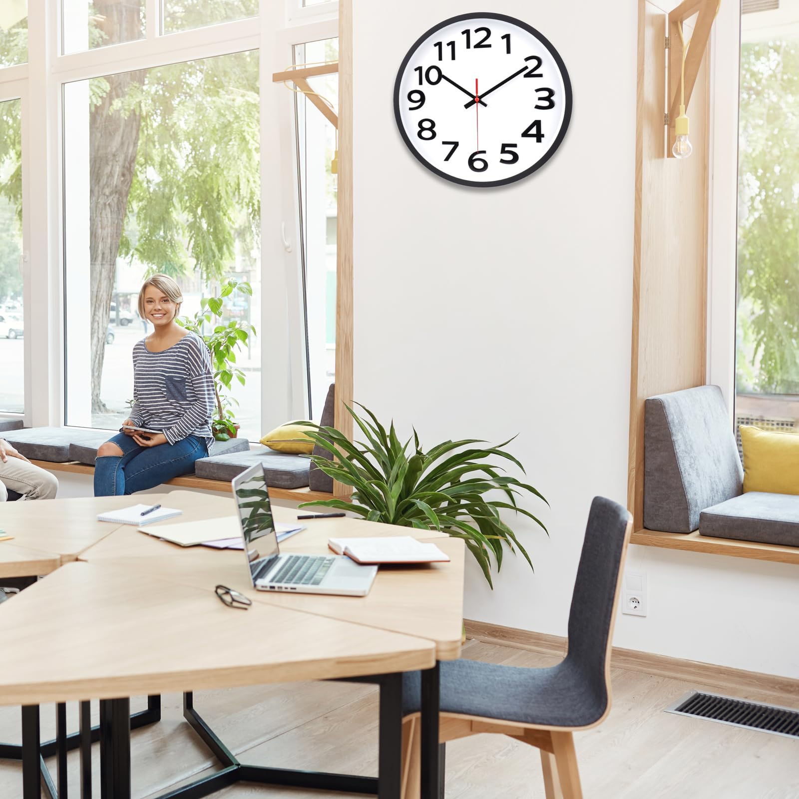 Uhr an einer weißen Wand. Eine Frau sitzt am Fenster. Tische, Pflanzen und ein Laptop befinden sich im Raum.