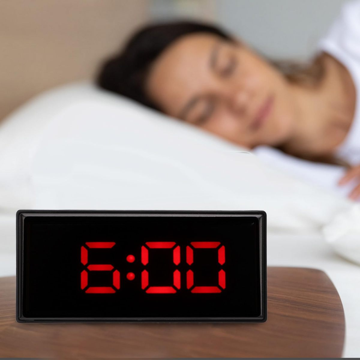 Una sveglia che mostra le 6:00 è appoggiata su una superficie di legno. Una donna dorme sullo sfondo su un cuscino e lenzuola bianche.