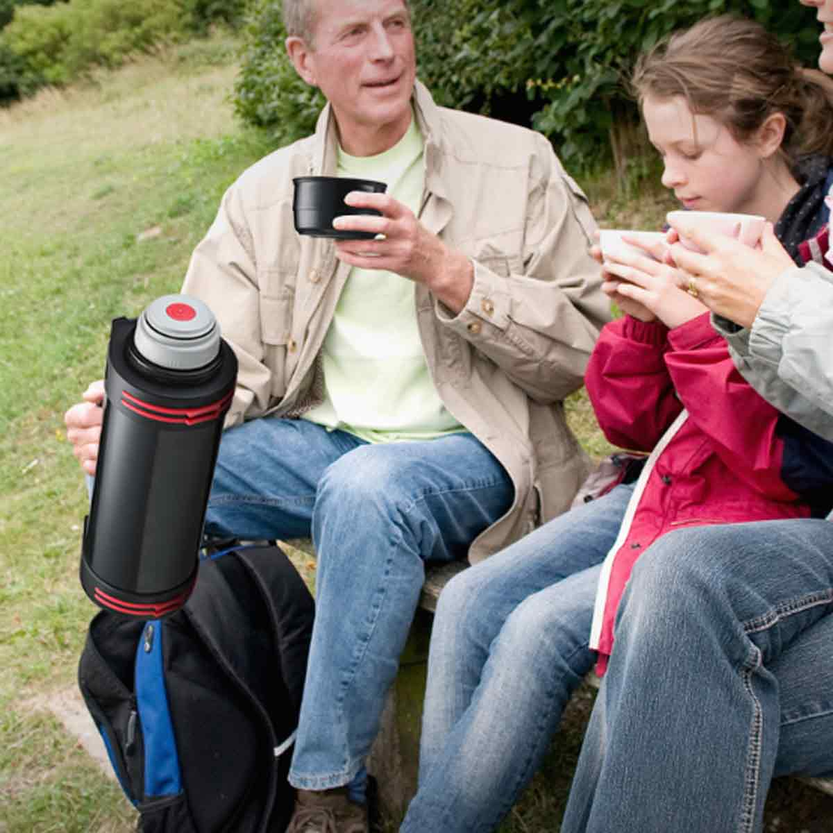 Eine Familie sitzt im Freien und trinkt aus Tassen. Eine Thermoskanne steht daneben. Der Mann hält eine schwarze Tasse. Die Frau und das Kind halten weiße Tassen.