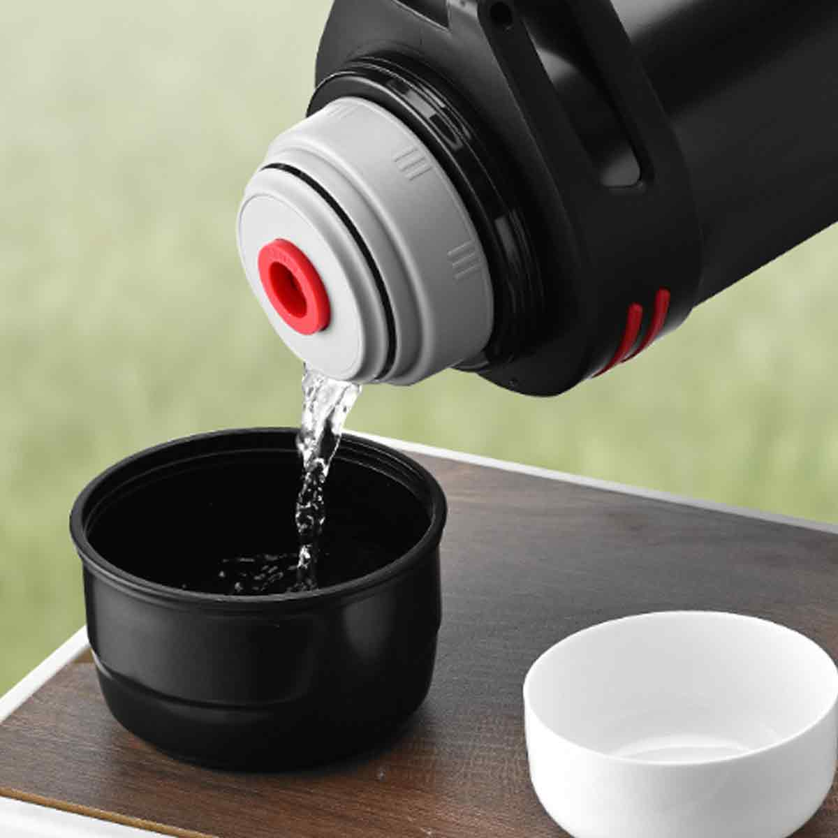 Eine schwarze Thermoskanne gießt Wasser in eine schwarze Tasse. Eine weiße Tasse steht daneben. Der Hintergrund ist unscharf grün.
