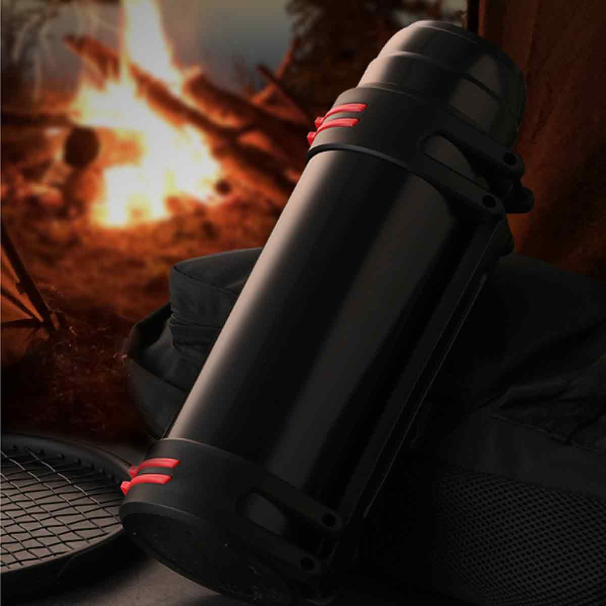 Eine schwarze Thermoskanne mit roten Akzenten vor einem Lagerfeuer. Ein Tennisschläger und ein dunkler Rucksack sind ebenfalls zu sehen.