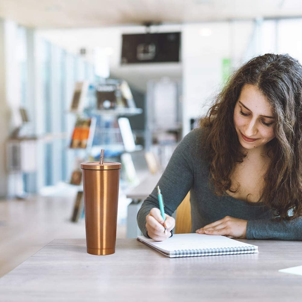 Eine Frau schreibt in einem Notizbuch an einem Schreibtisch, daneben steht ein kupferfarbener Becher mit Strohhalm. Im Hintergrund sind Bücherregale und ein Bildschirm zu sehen.