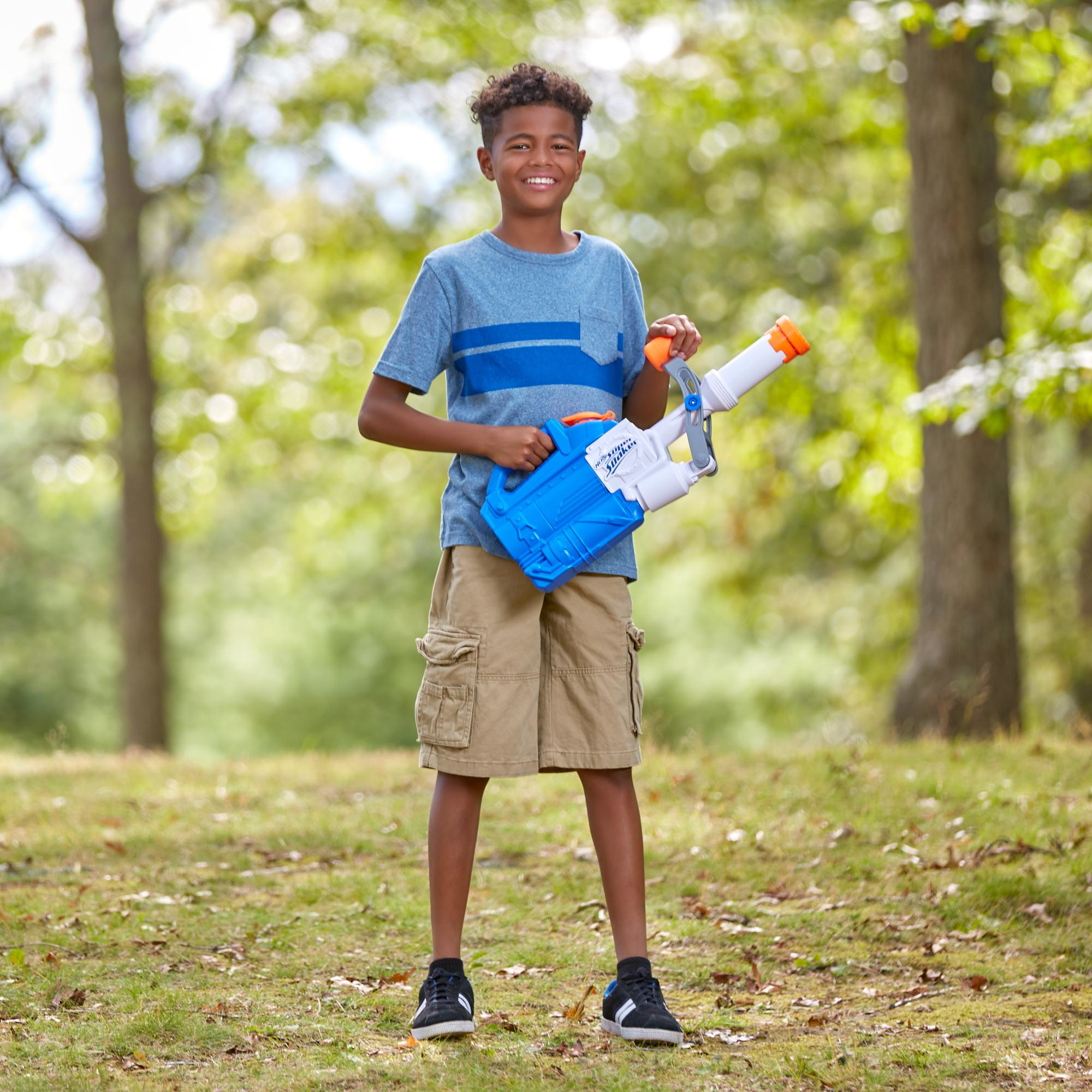 Un ragazzo sorridente tiene in mano una pistola ad acqua blu e bianca in un parco. Alberi ed erba sullo sfondo.