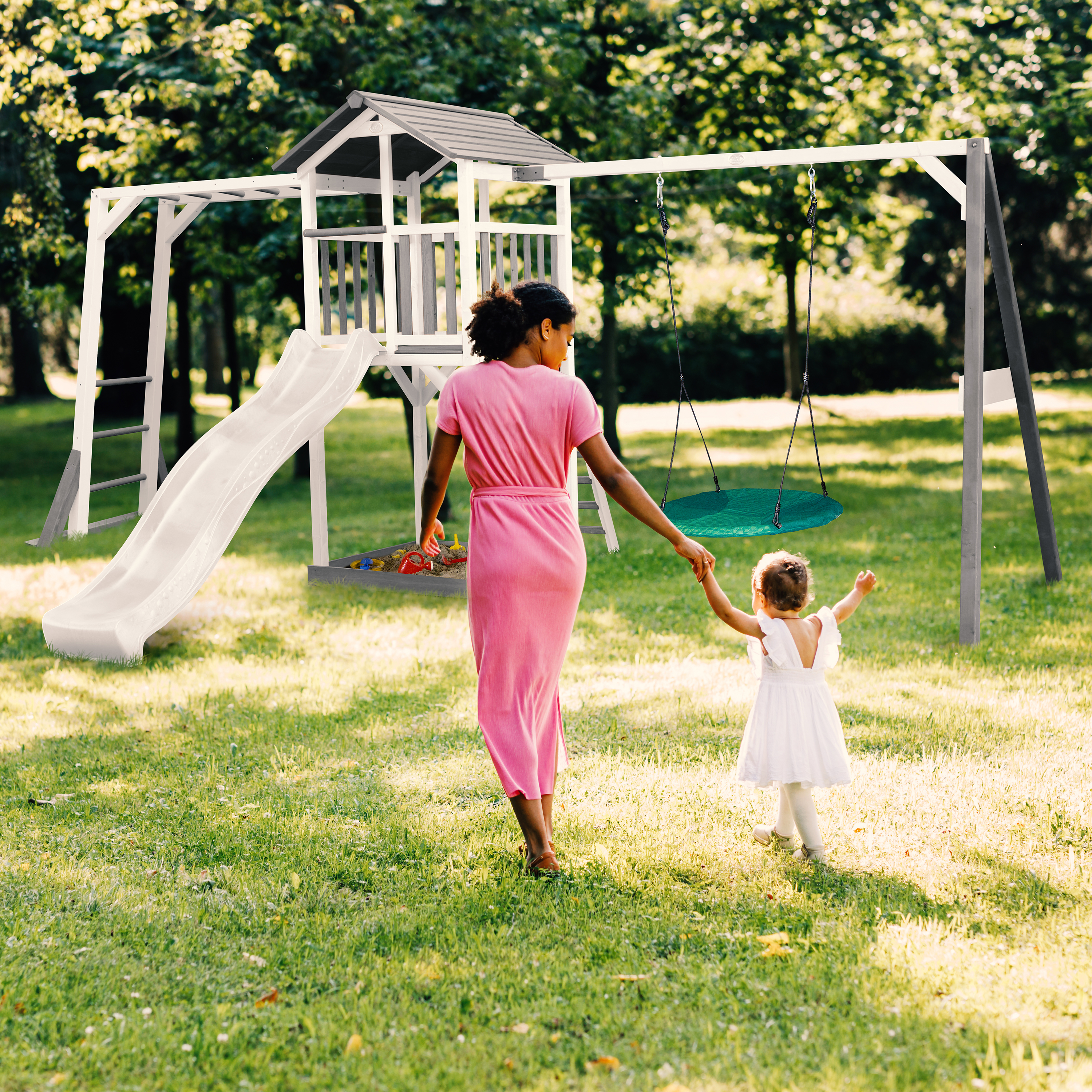 Eine Frau und ein Kind gehen auf einen Spielplatz zu. Im Hintergrund befinden sich eine Schaukel und eine Rutsche.
