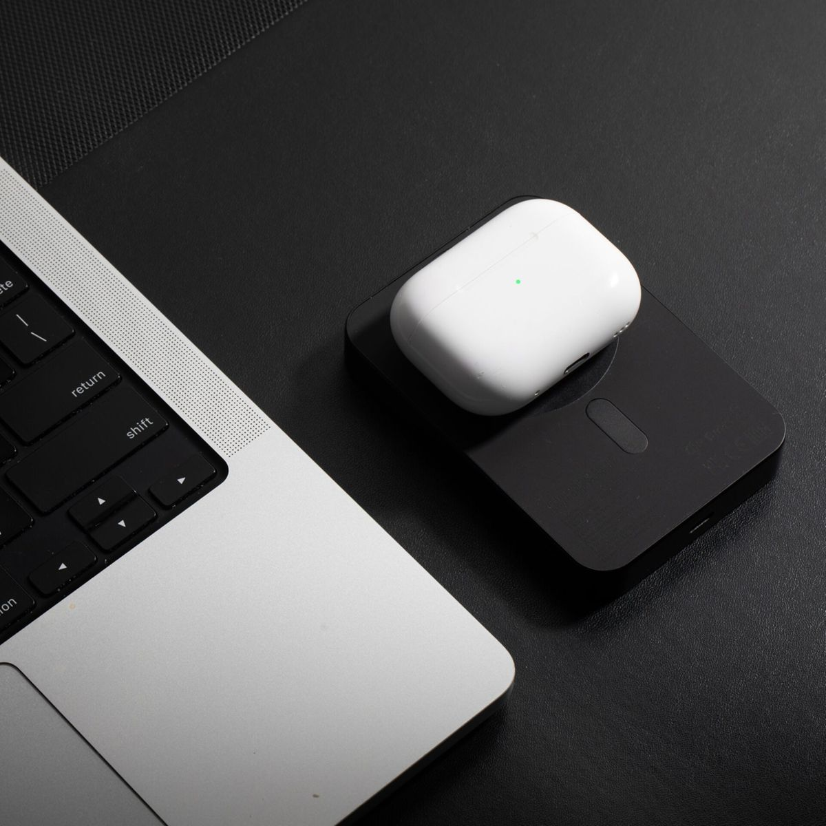 Una custodia di ricarica bianca per AirPods è appoggiata su un caricatore nero, accanto a un laptop argentato su una superficie nera.