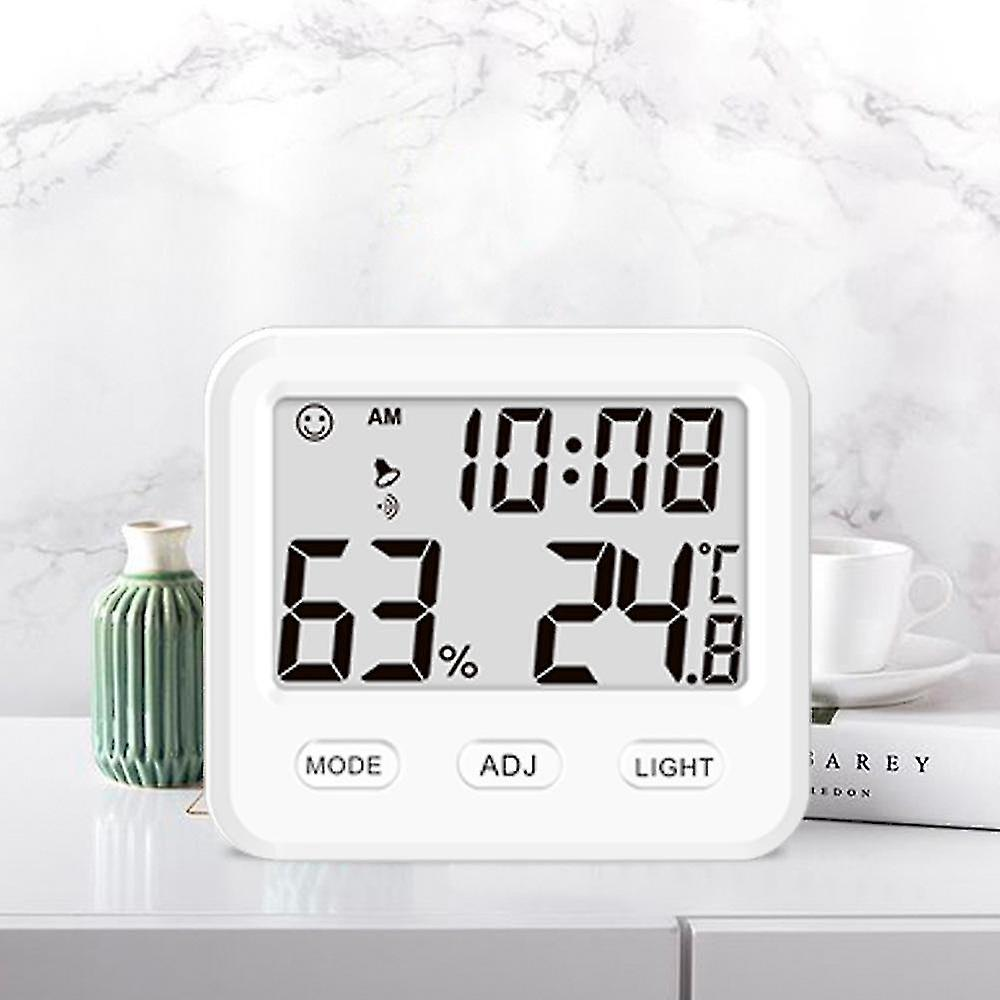 Eine weiße Digitaluhr zeigt Zeit, Luftfeuchtigkeit und Temperatur an. Eine grüne Vase, ein Buch und eine Teetasse stehen daneben auf einer weißen Oberfläche.