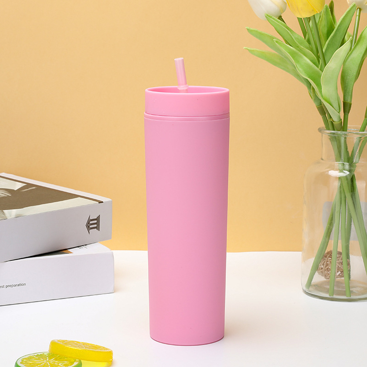 Rosa Becher mit Strohhalm auf weißer Oberfläche, mit Blumenvase und Büchern im Hintergrund. Zitronenscheiben im Vordergrund.