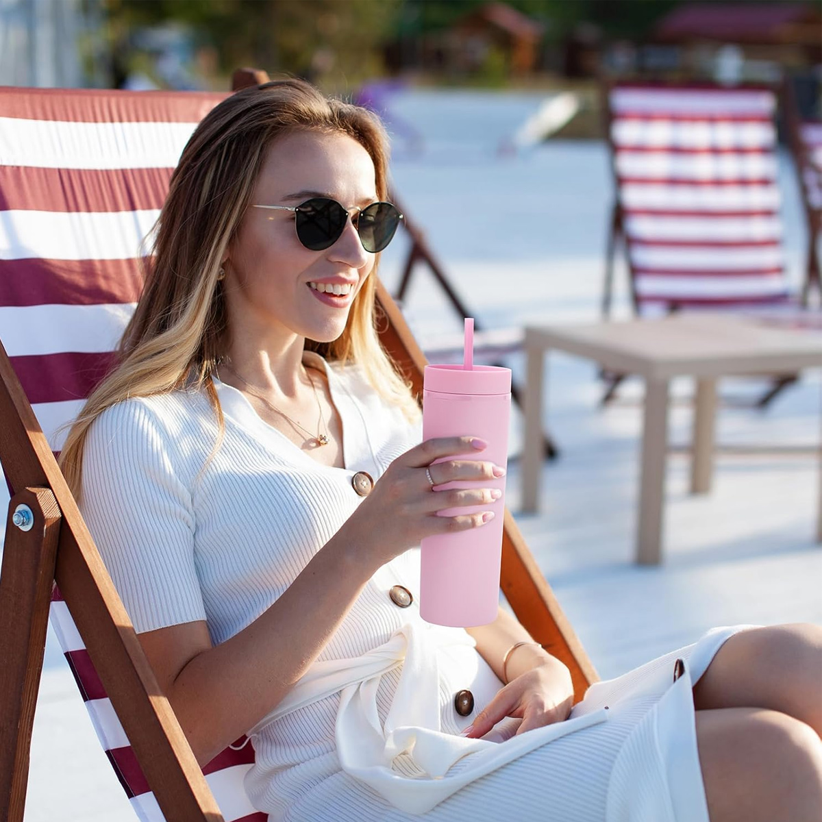 Frau mit Sonnenbrille lächelt, hält rosa Becher, sitzt auf Strandstuhl. Hintergrund Strand, Tische und Stühle.